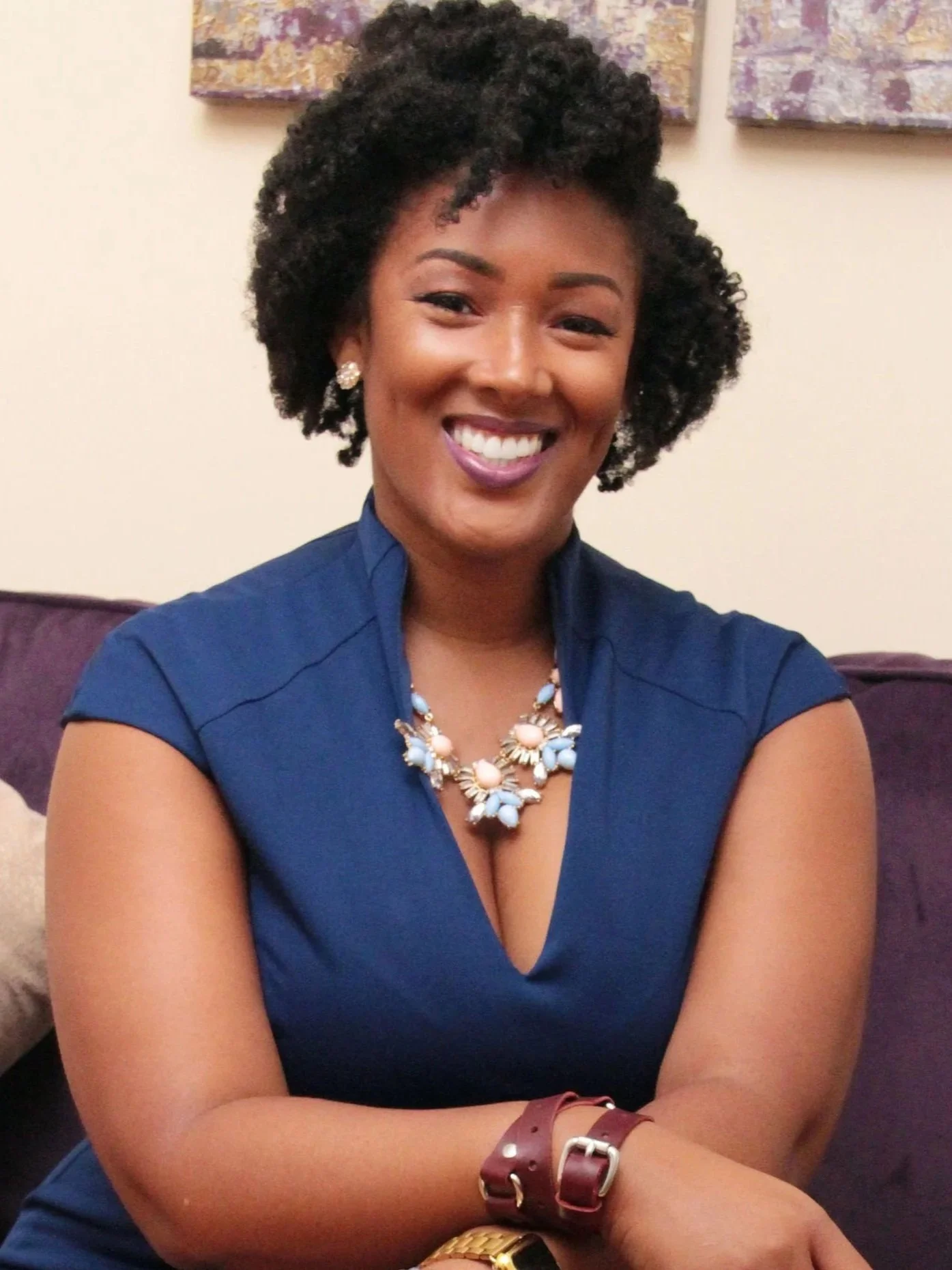A smiling woman with short curly hair sitting on a purple couch, wearing a blue dress and jewelry.