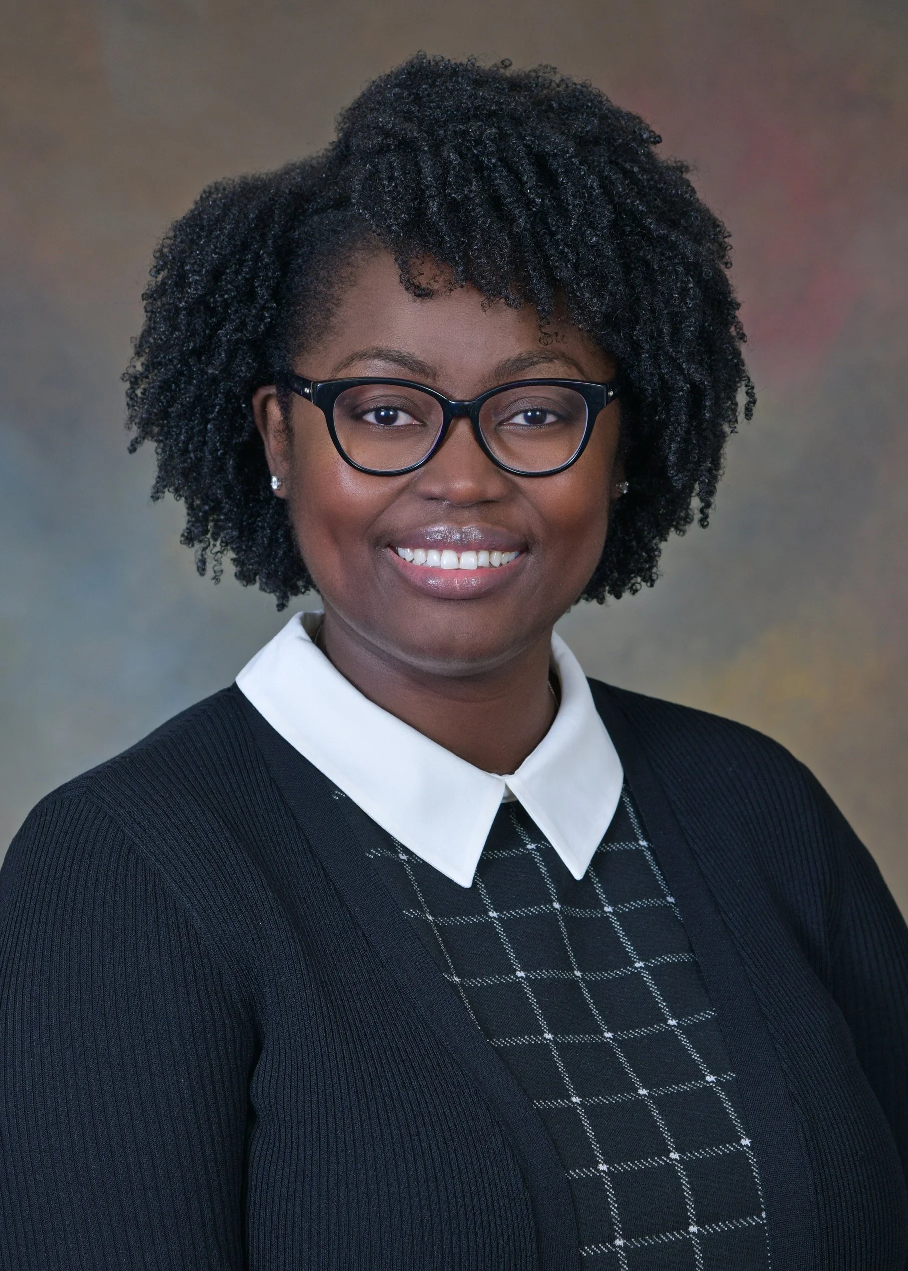 A woman with dark curly hair, glasses, and earrings, smiling in a professional portrait with a blurred multicolored background.