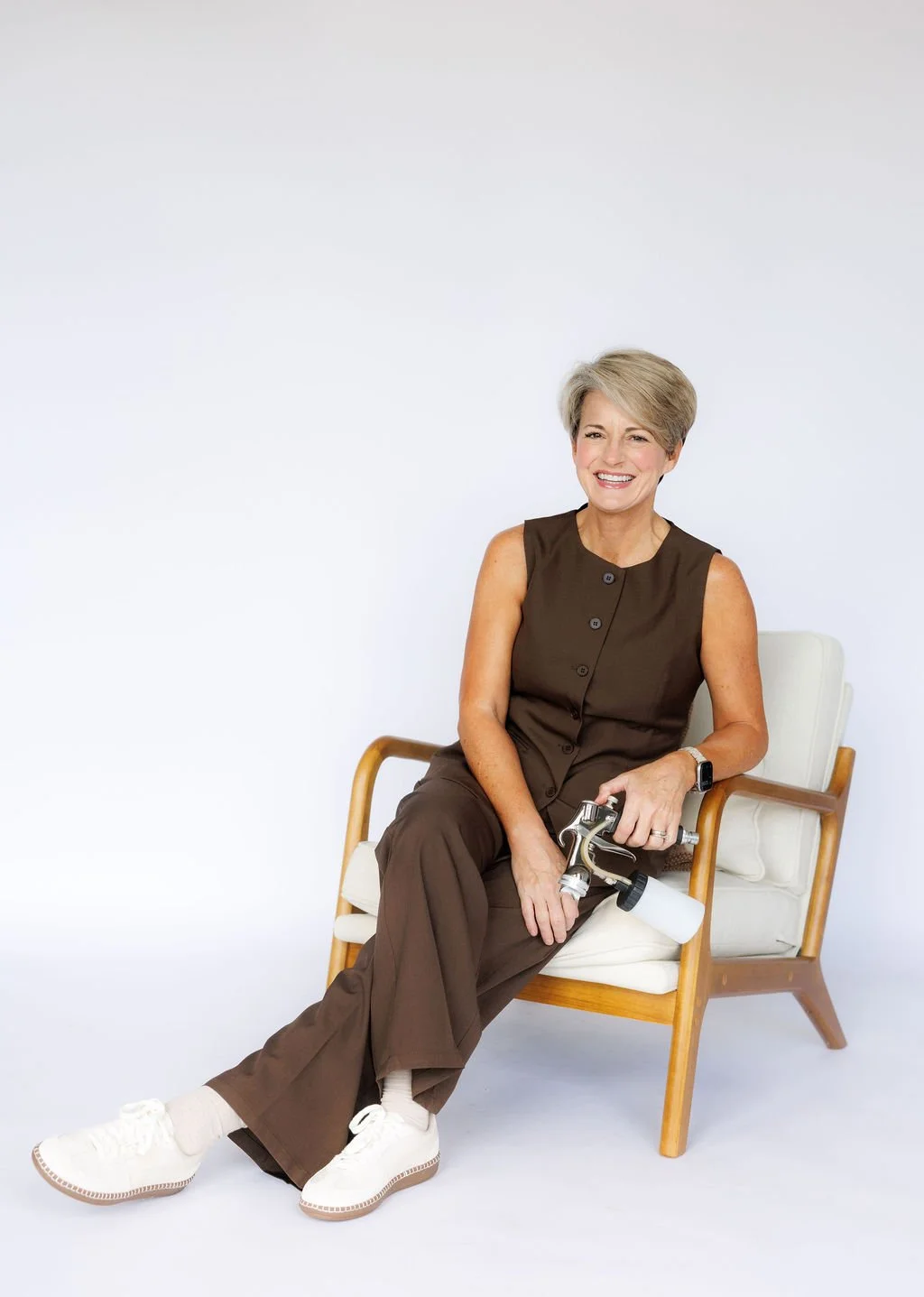 A woman with short blonde hair smiling, sitting on a modern wooden chair with white cushion, wearing a sleeveless brown outfit, white sneakers, a watch, and spray tan equipment.