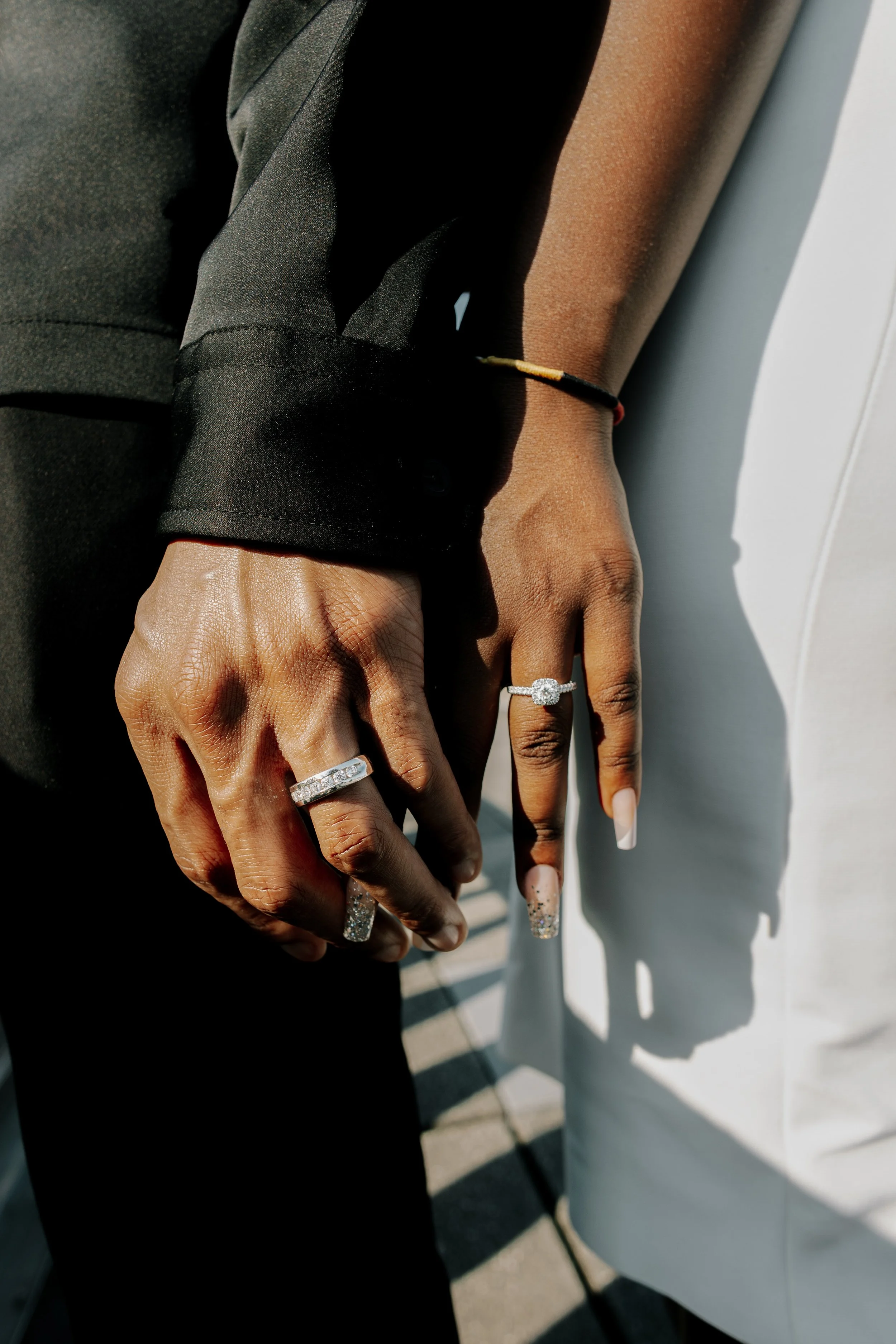 Close-up of two people's hands clasped together, showing rings on their fingers, with one person wearing a black suit and the other wearing a white outfit.