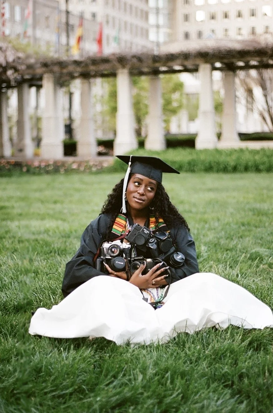 A young woman in graduation cap and gown sitting on grass holding multiple cameras while looking up thoughtfully, with a green park and a building with columns in the background.