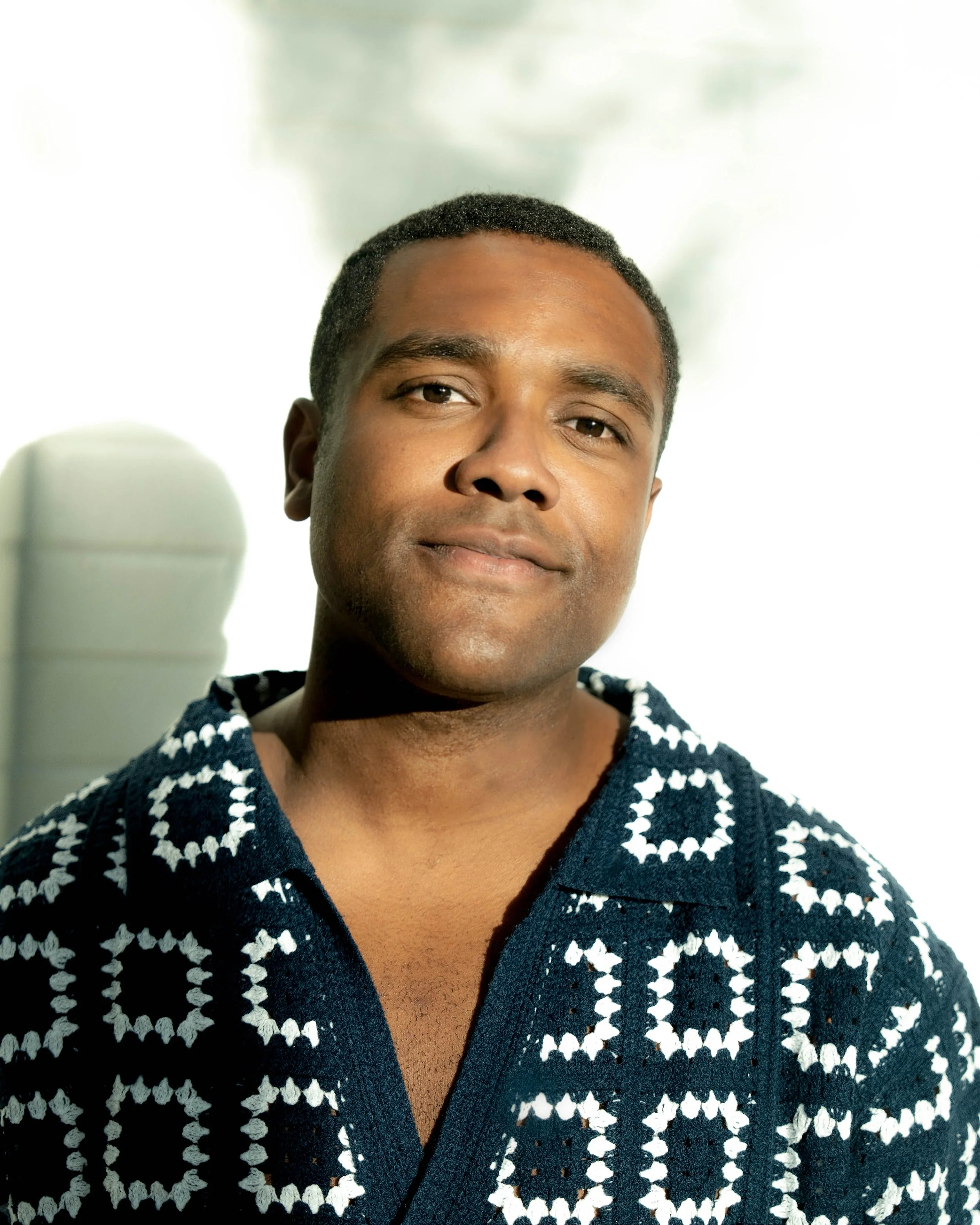 Portrait of a young man with short hair, wearing a navy blue and white patterned shirt, smiling softly, with a blurred outdoor background.