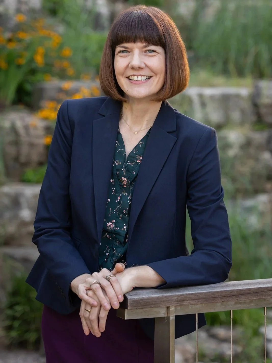 Emily Grannemann with short brown hair, wearing a navy blazer and floral blouse, leaning on a wooden railing outdoors, smiling at the camera with a blurred garden background.