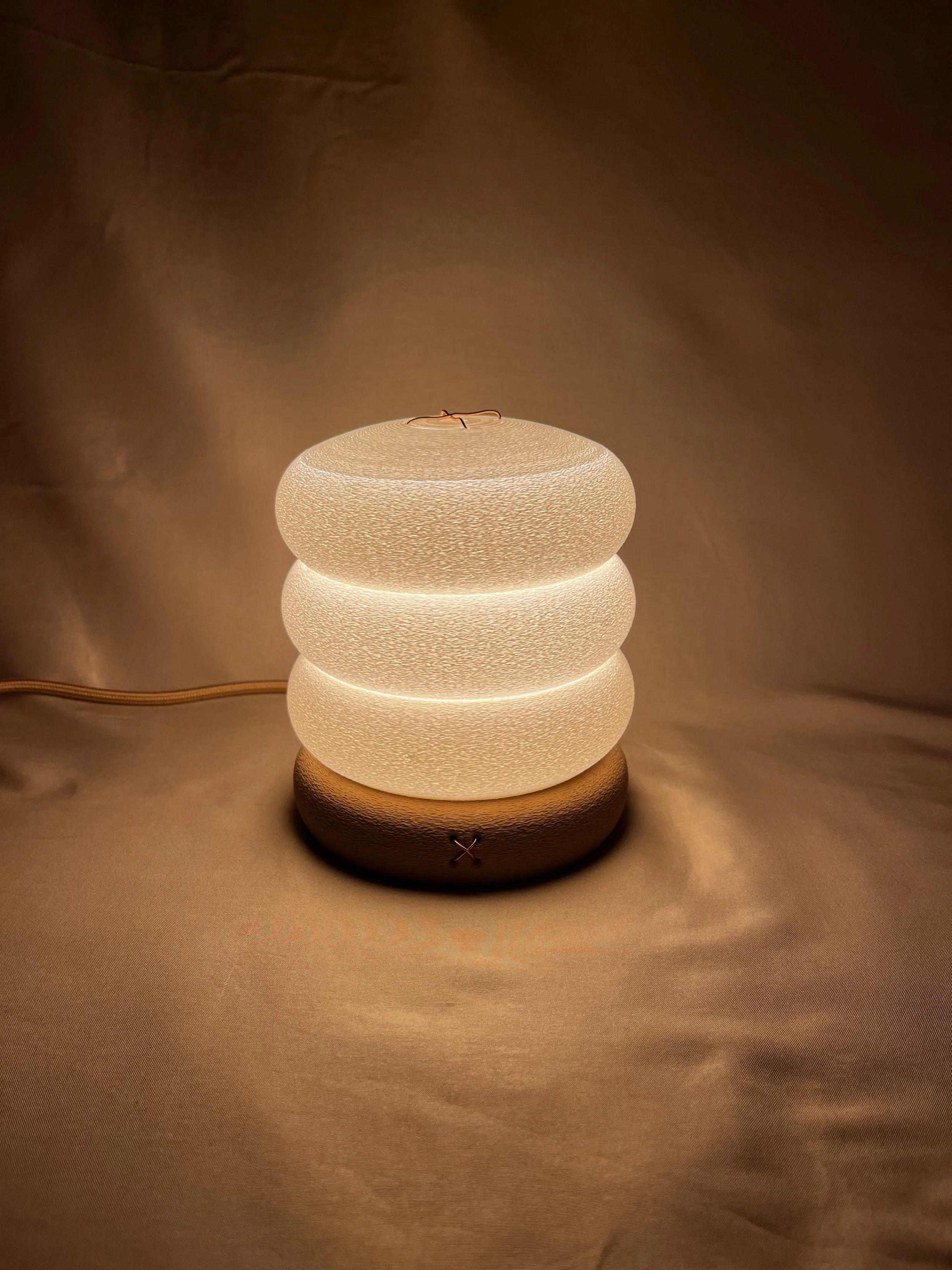 A modern table lamp with a textured, round, layered design and warm lighting on a soft fabric surface.