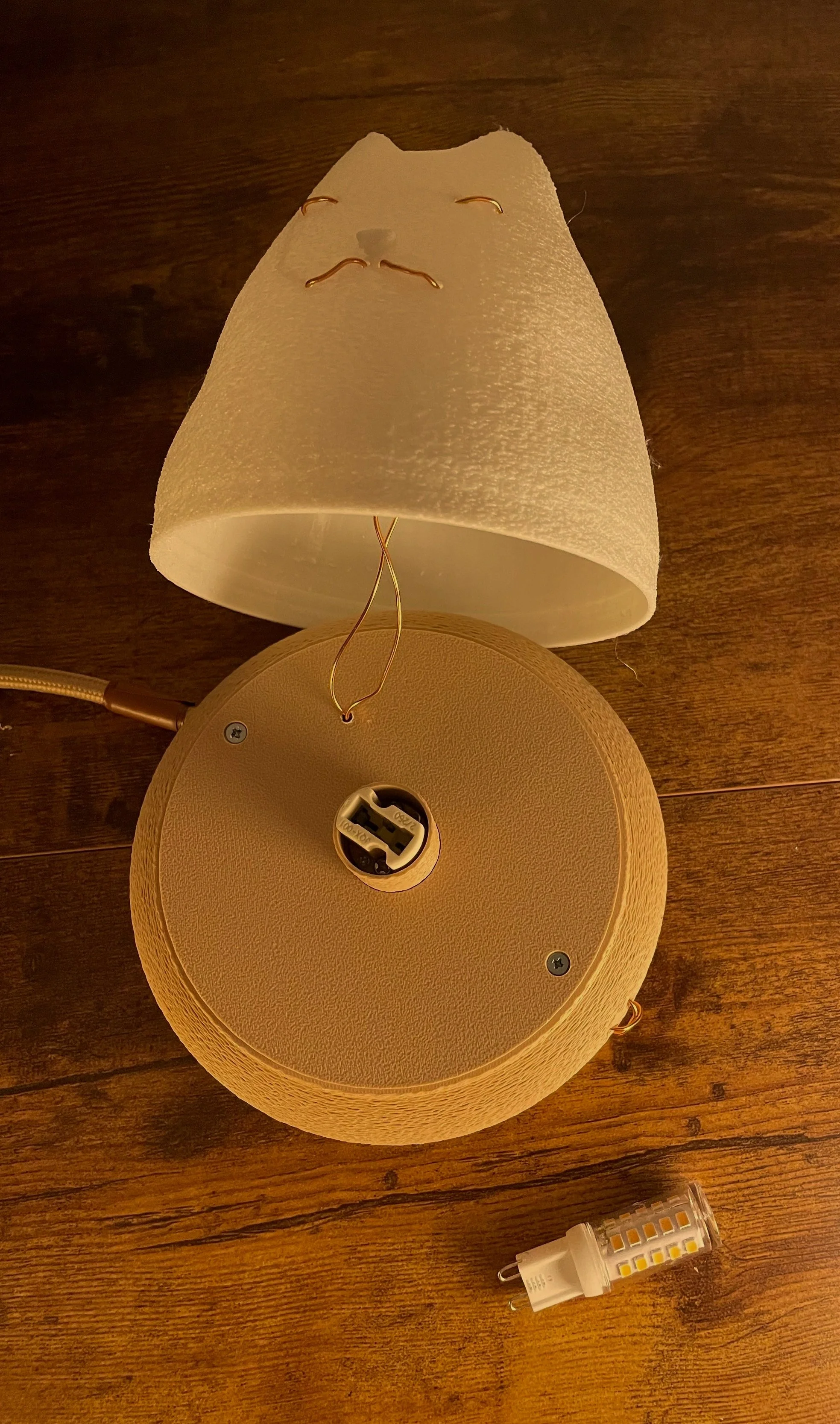 The image shows a disassembled table lamp with a beige textured base, a white lampshade with a face drawn on it, and a small LED light bulb placed on a wooden surface.