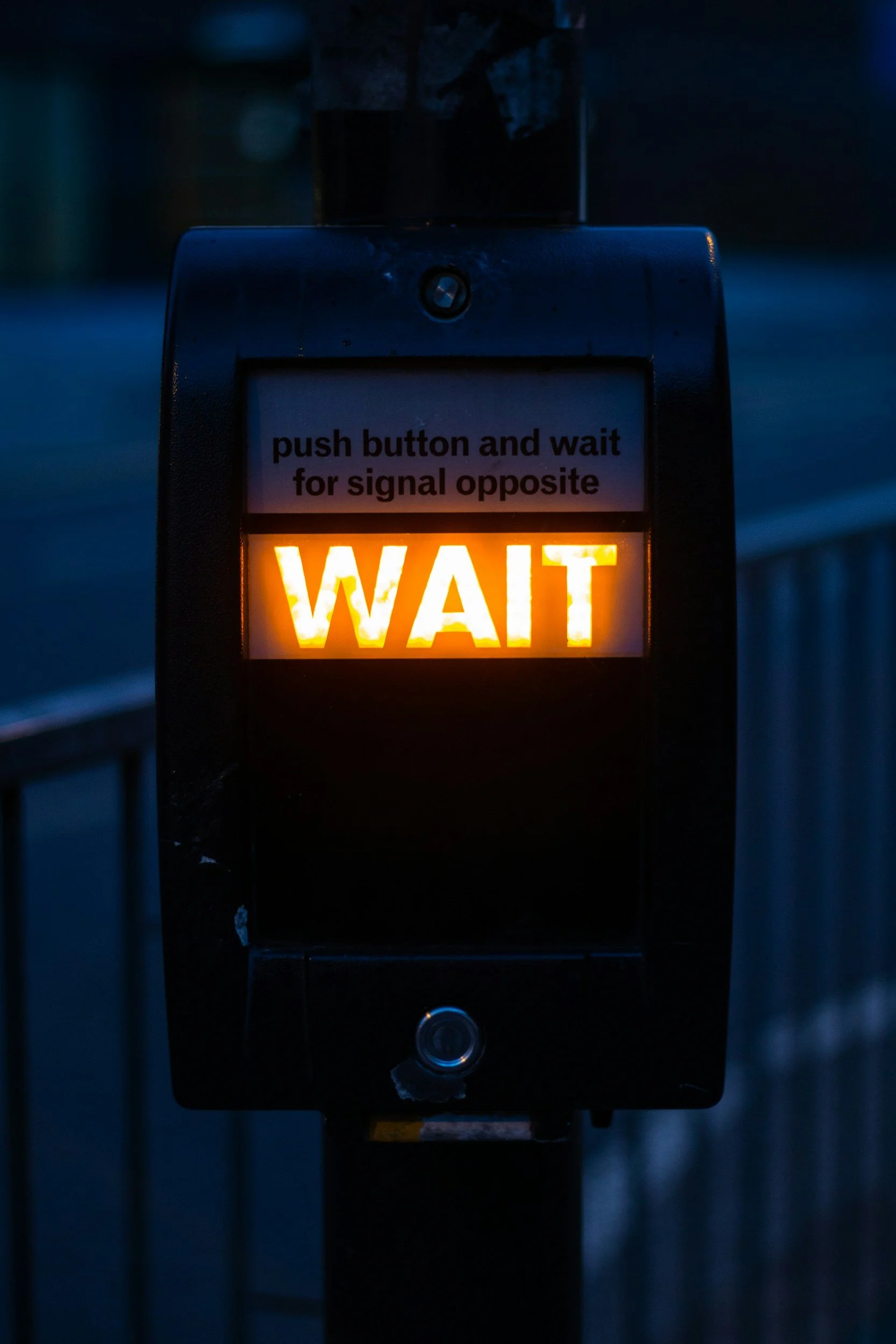 A close-up view of a pedestrian crossing button with a digital display that says "WAIT" in bright orange lights. Above the display, a sign instructs to "push button and wait for signal opposite." The background is dark, suggesting it's evening or early morning.