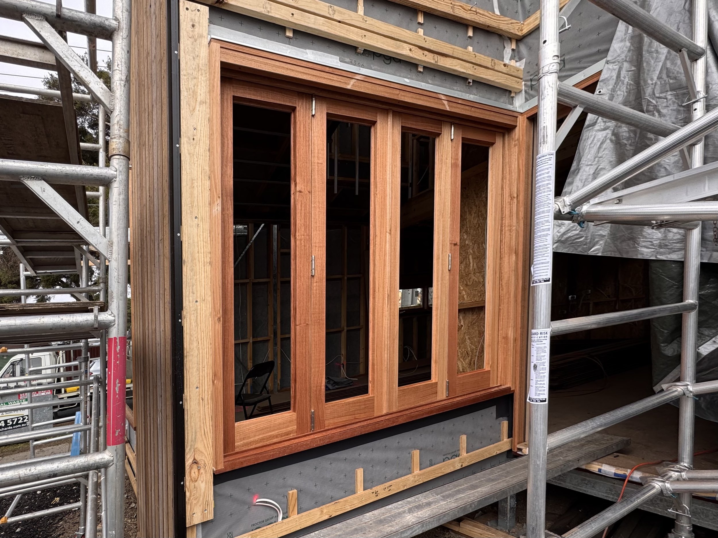 Construction site showing a small wooden framed structure with glass doors, surrounded by scaffolding.