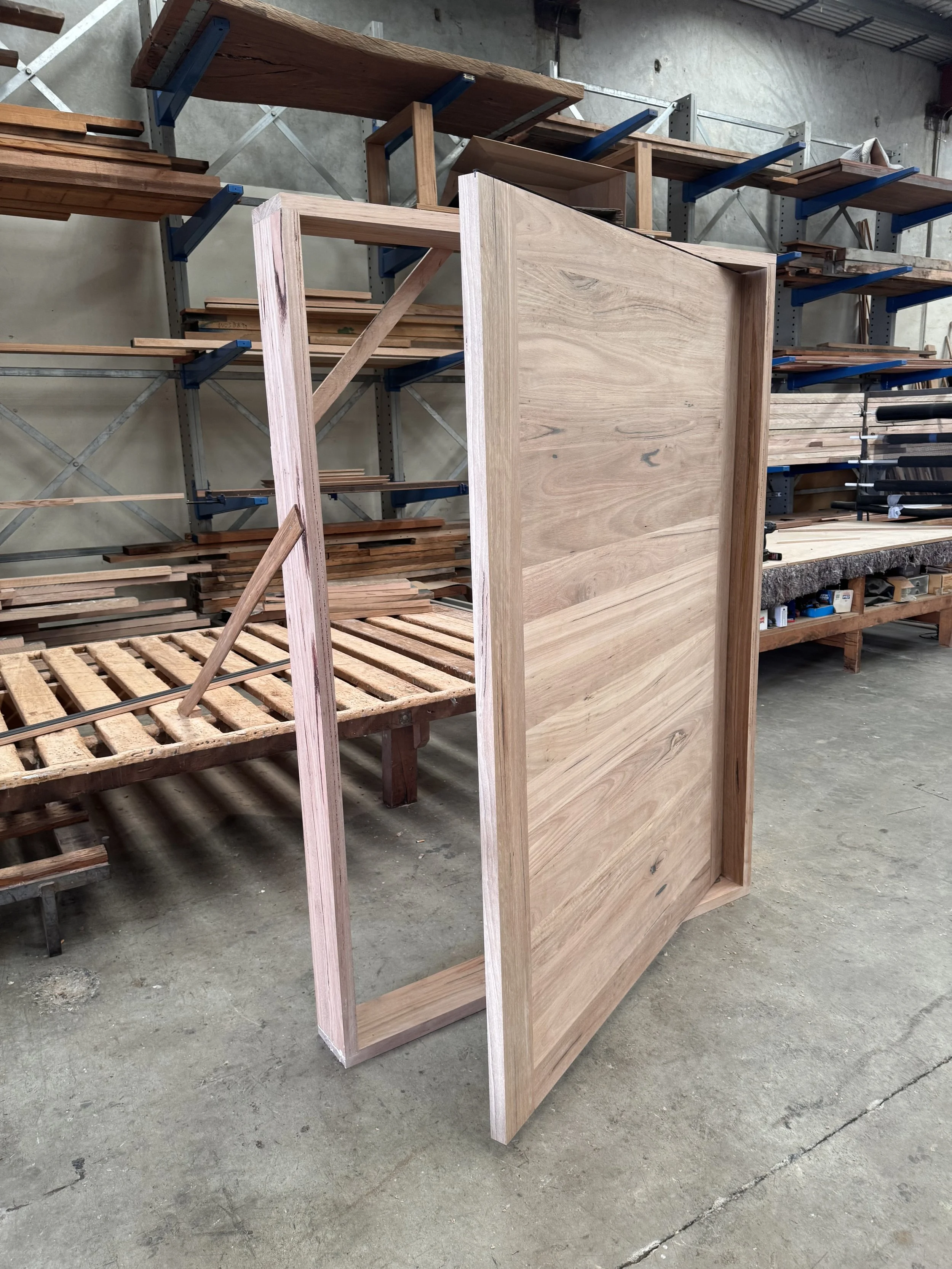 Tasmanian Oak Pivot door in a woodworking shop, surrounded by shelves with wood planks and tools.