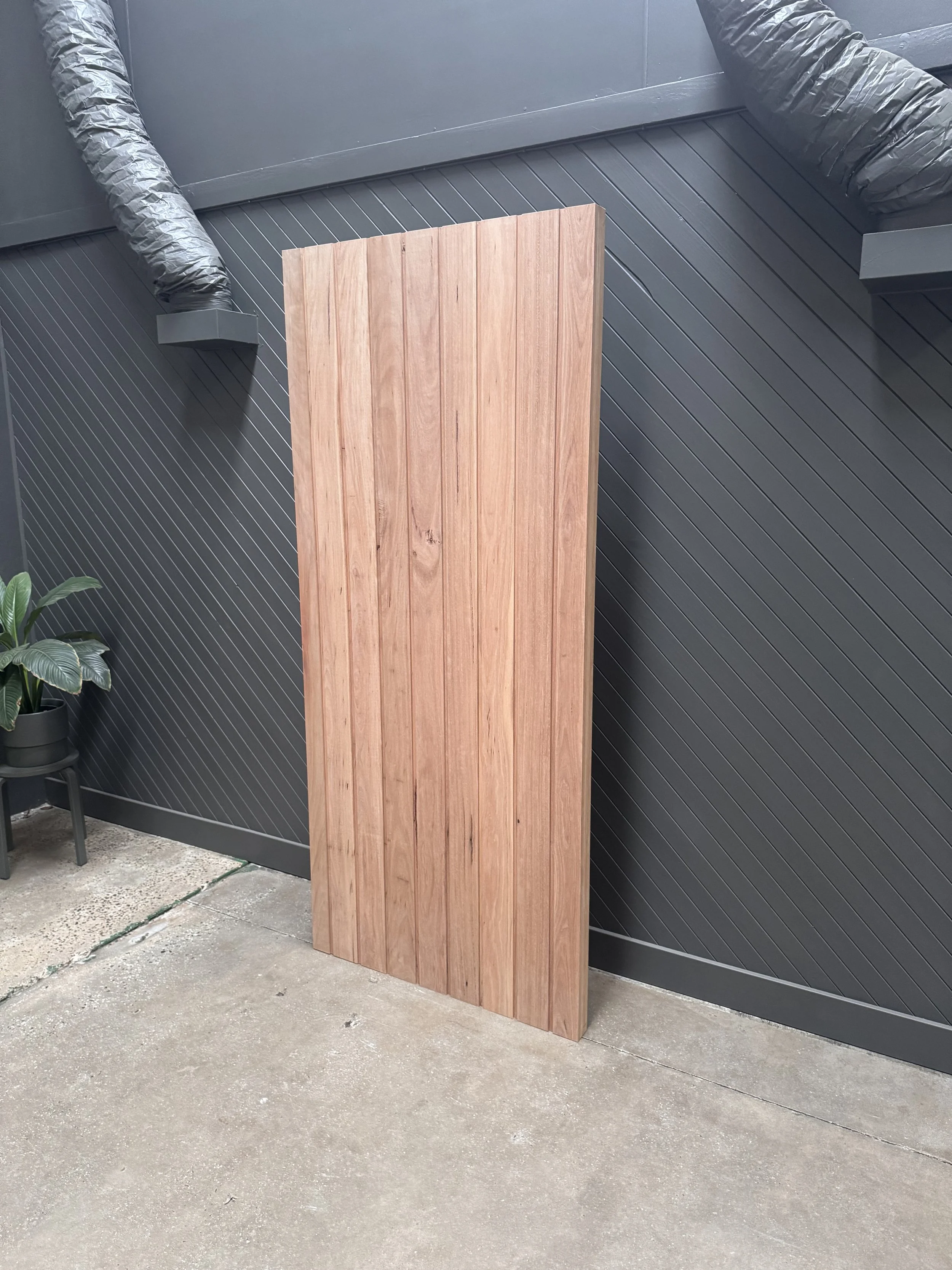 A tall wooden plank leaning against a dark gray wall with diagonal paneling, near a concrete floor and a potted plant.