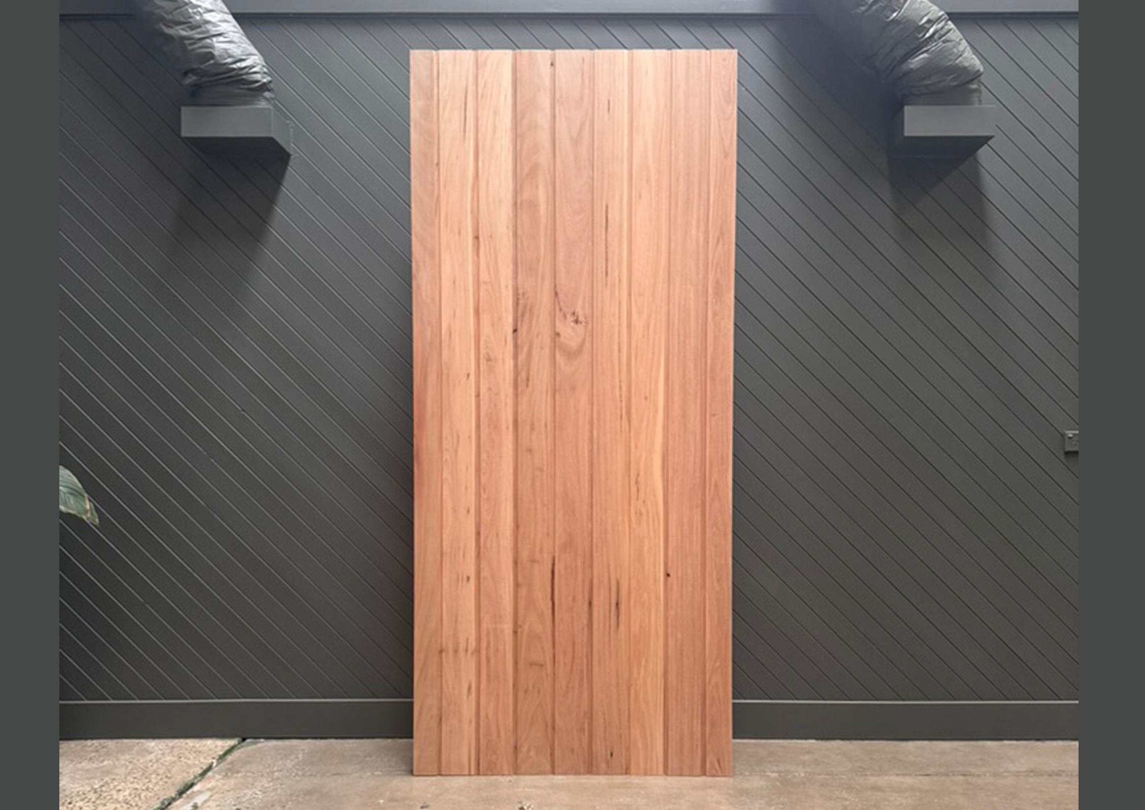 Wooden door or panel leaning against a dark gray wall with diagonal siding, with black exhaust vent pipes on either side.