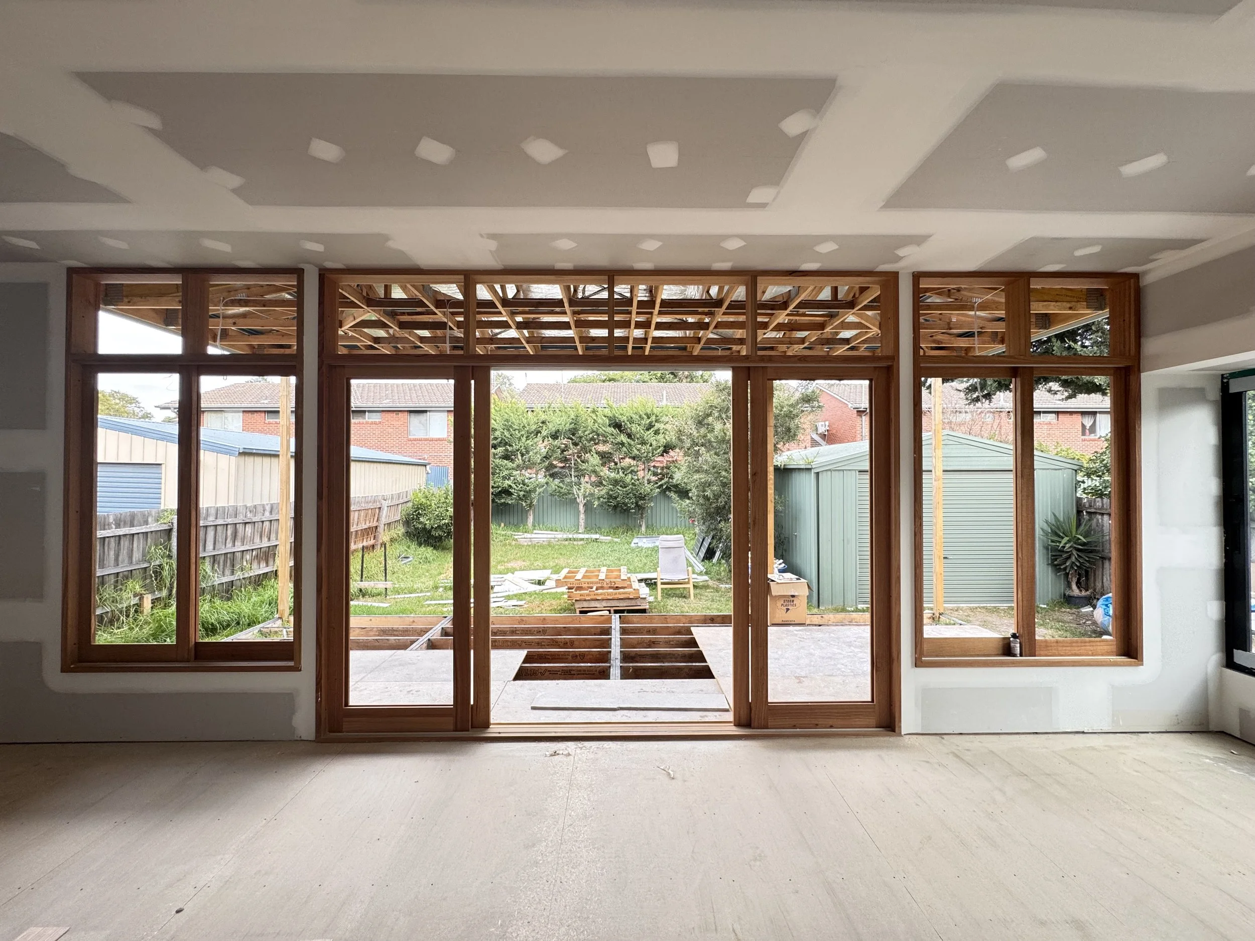 Interior view of a room under construction with large wooden-framed windows and glass doors overlooking a backyard with a green shed, trees, and a grassy lawn.