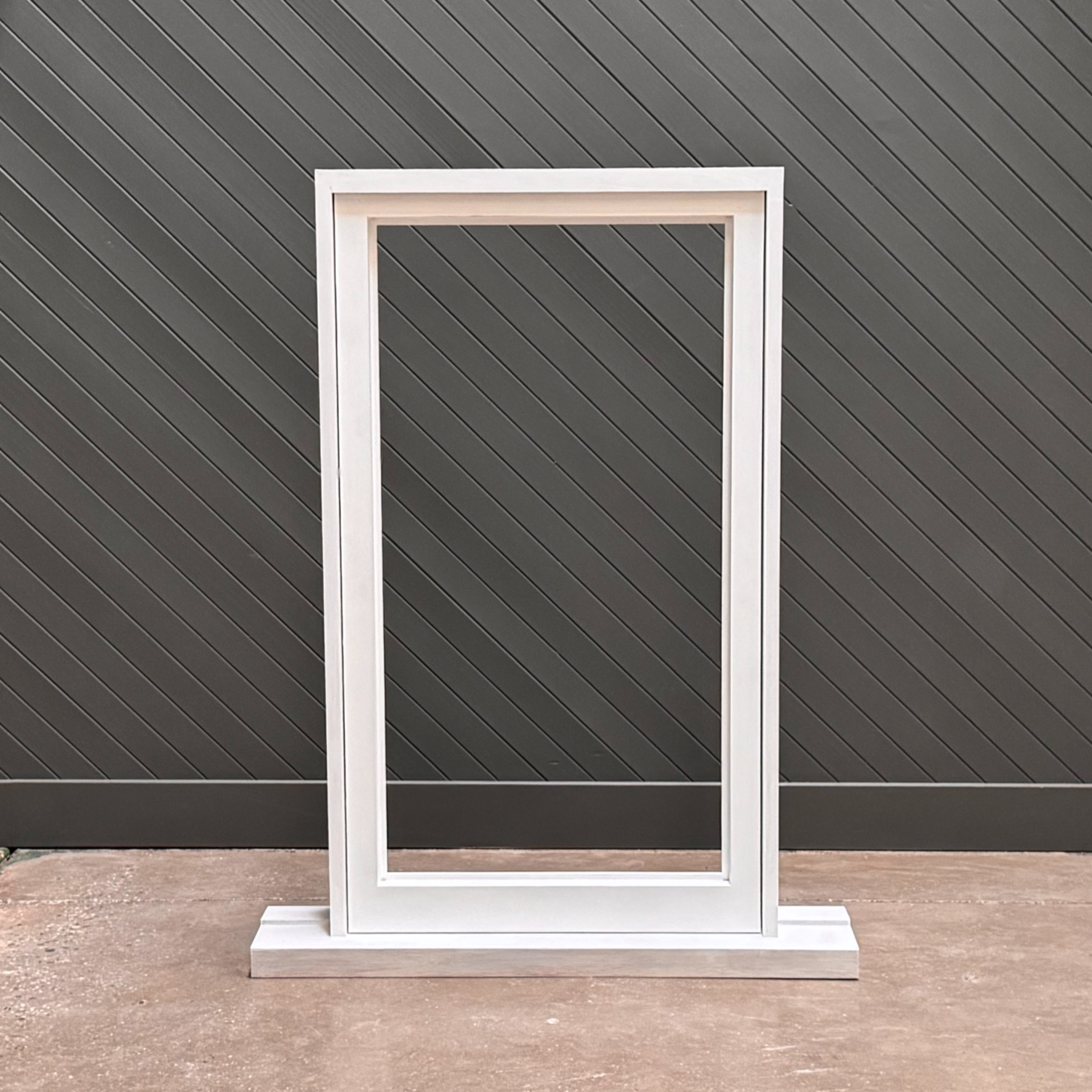 Empty white picture frame standing on a concrete ground against a dark gray, diagonally striped wall.