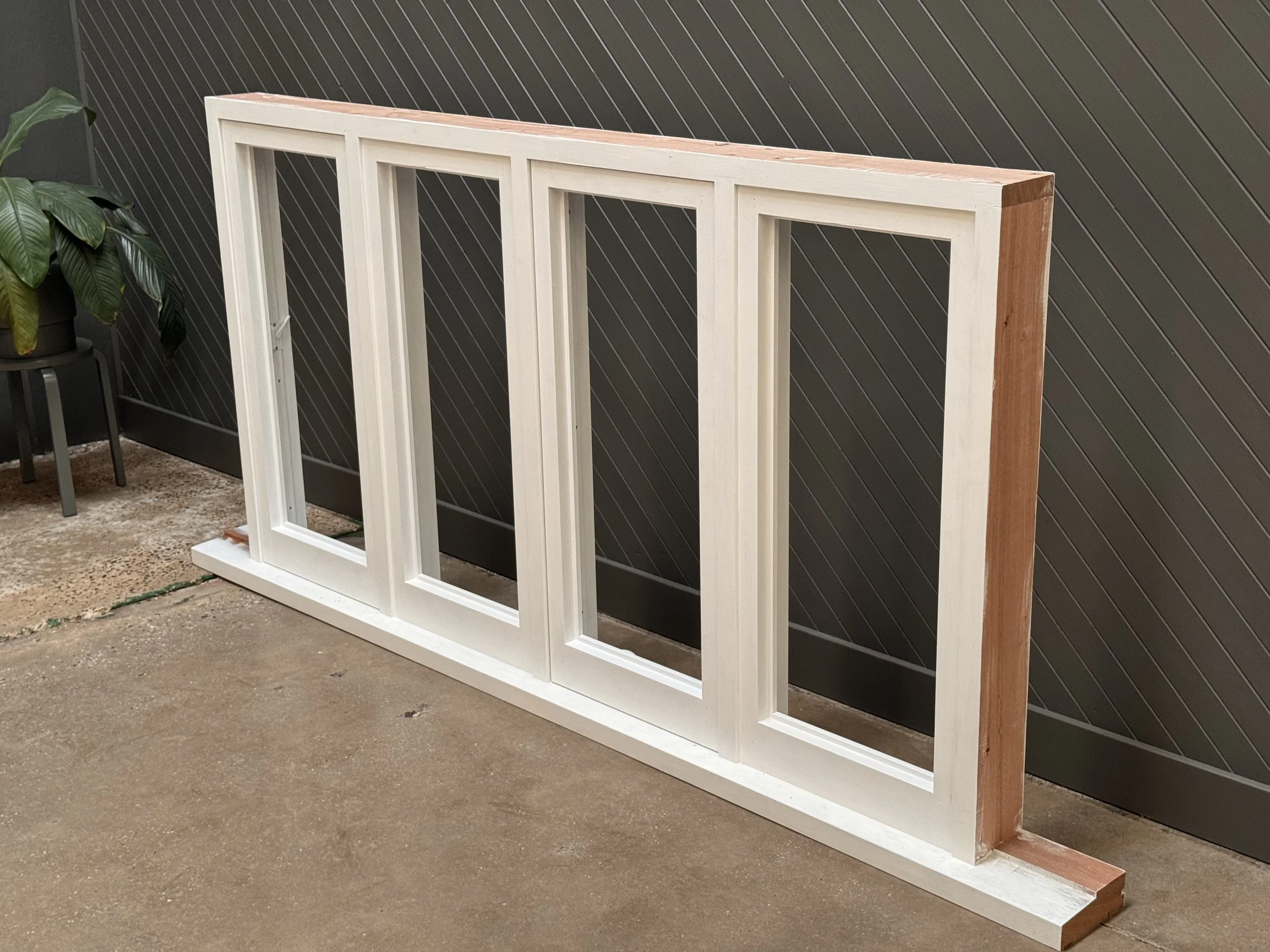 Wooden window frame with four window openings leaning against a dark wall, next to a potted plant, on a concrete floor.