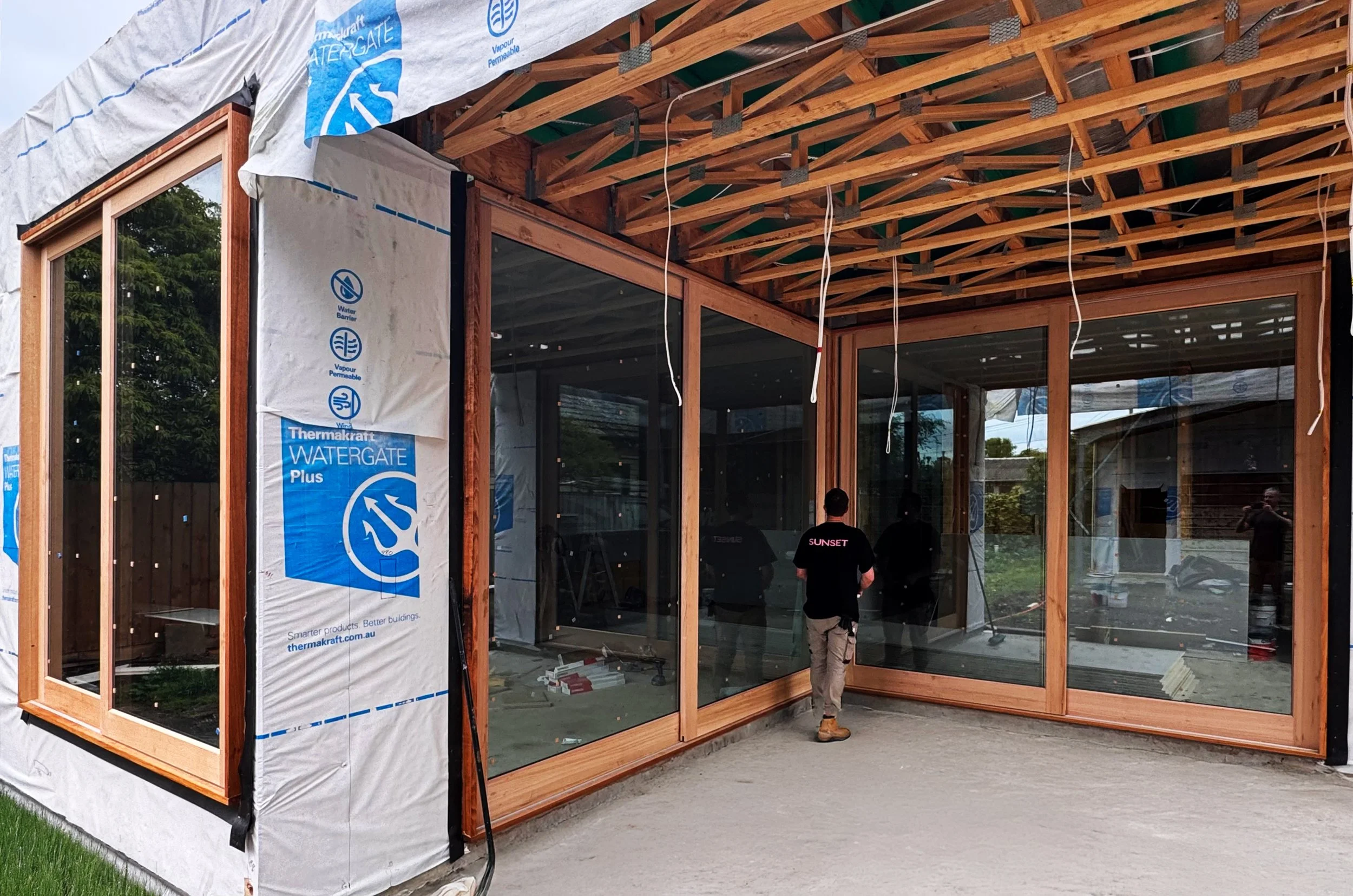 Construction site of a house with wooden framing and large glass windows, some insulation material visible on the exterior, and workers inside.