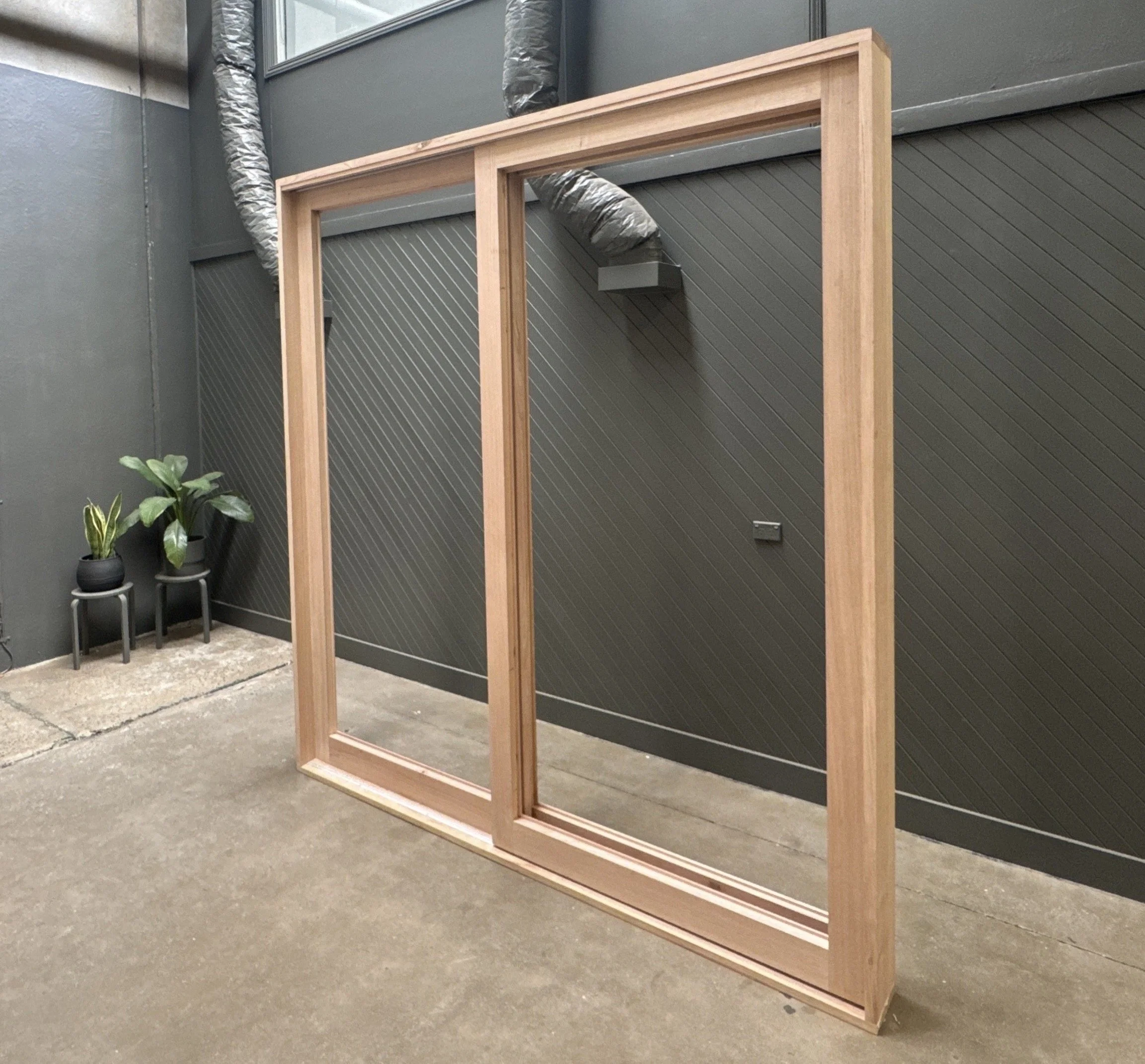 A wooden picture frame leaning against a dark gray wall in an indoor space, with two small potted plants on black stands in the corner on the left side of the image.