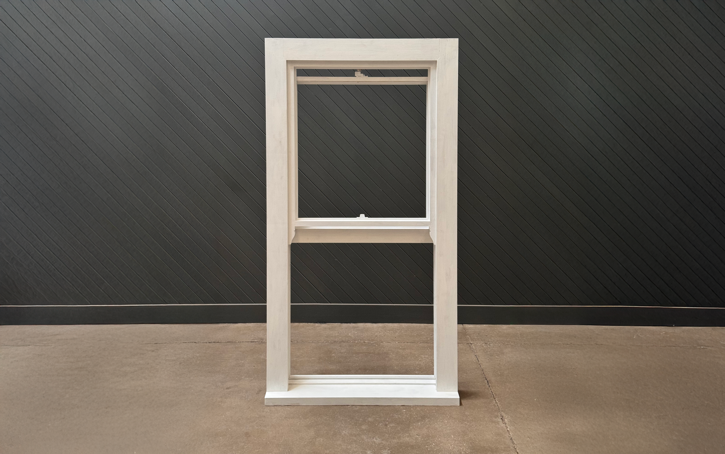 A white wooden picture frame standing on a concrete floor against a black textured wall.