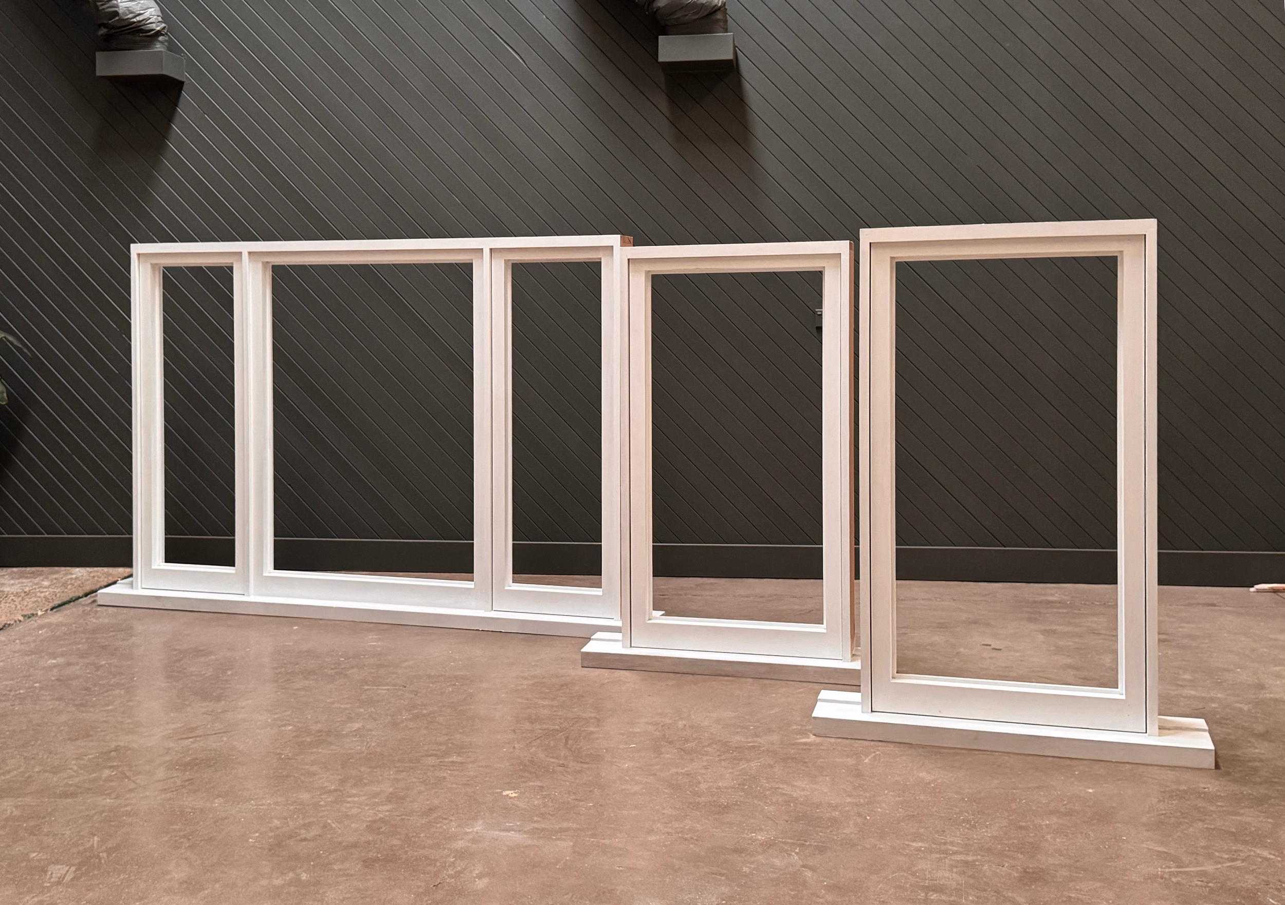 Four white window frames are lined up indoors against a dark wall with diagonal paneling, on a concrete floor.