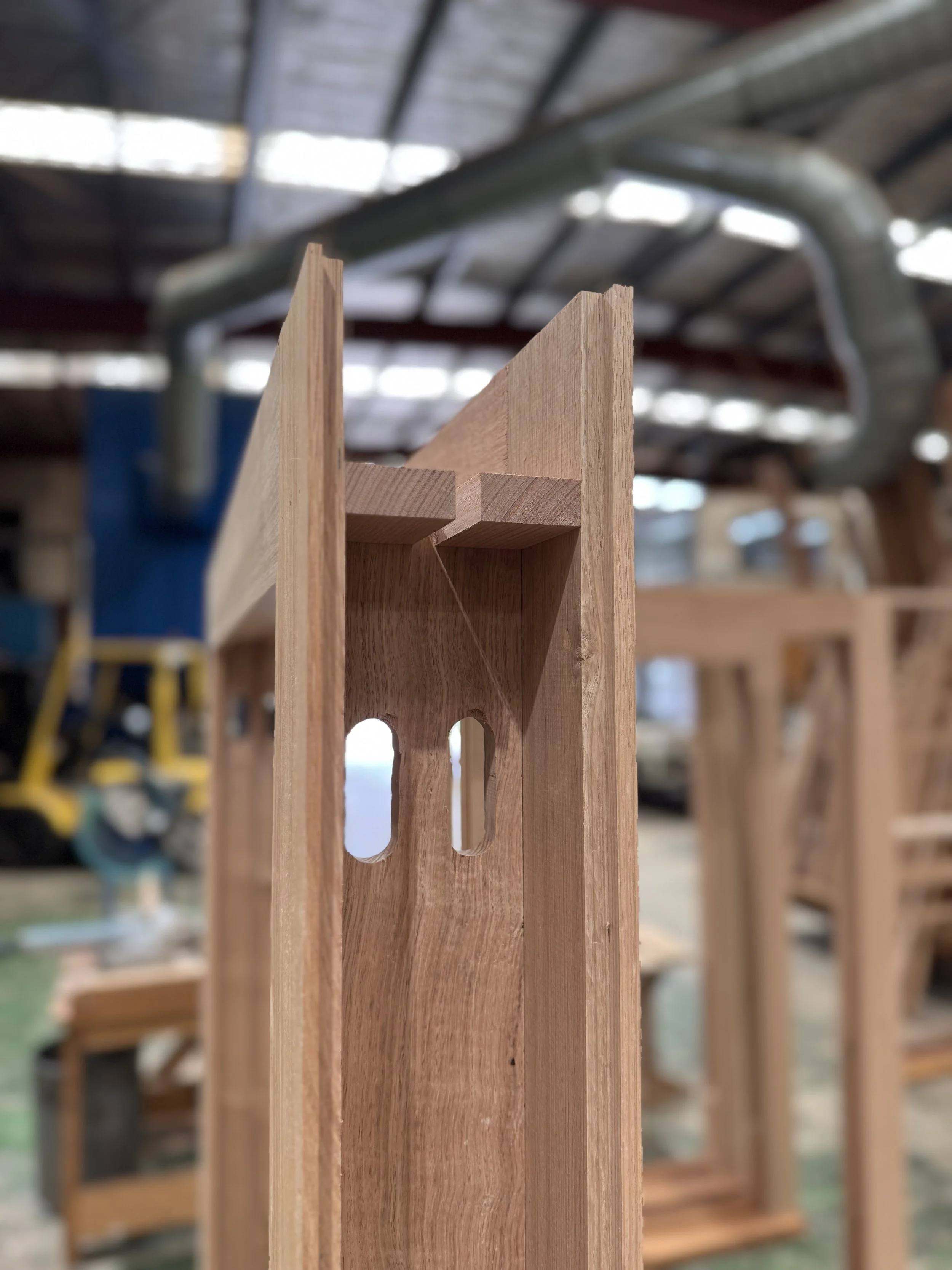 Close-up of a wooden furniture piece under construction in a workshop, with visible slots and joints, and blurred woodworking tools and equipment in the background.