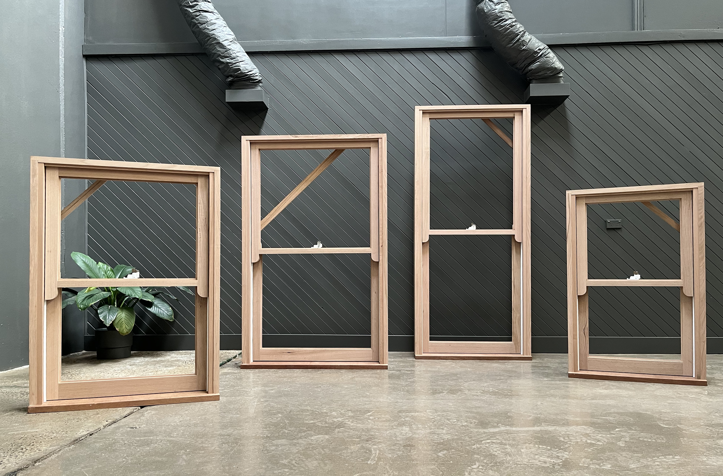 Four wooden picture frames of various sizes leaning against a black wall with ductwork. A potted green plant is in the corner on the left side of the image.