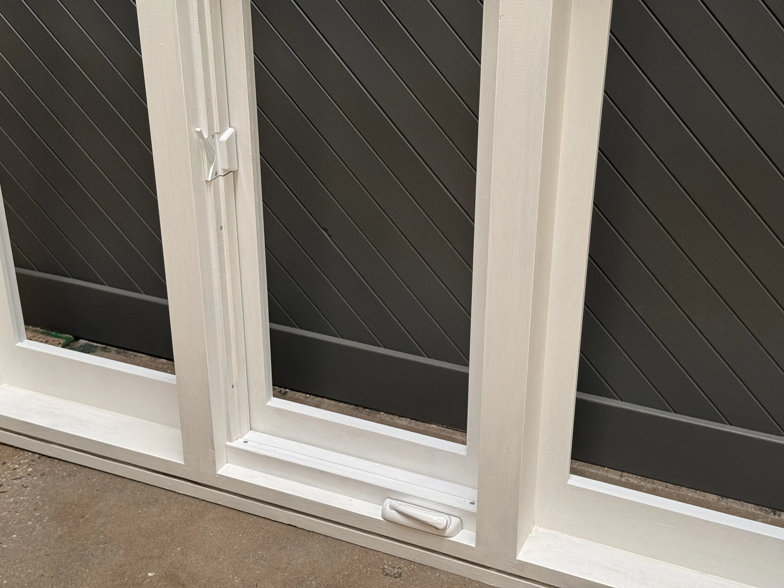 A white screen door with a latch and a mesh panel, in front of a black fence with diagonal siding.