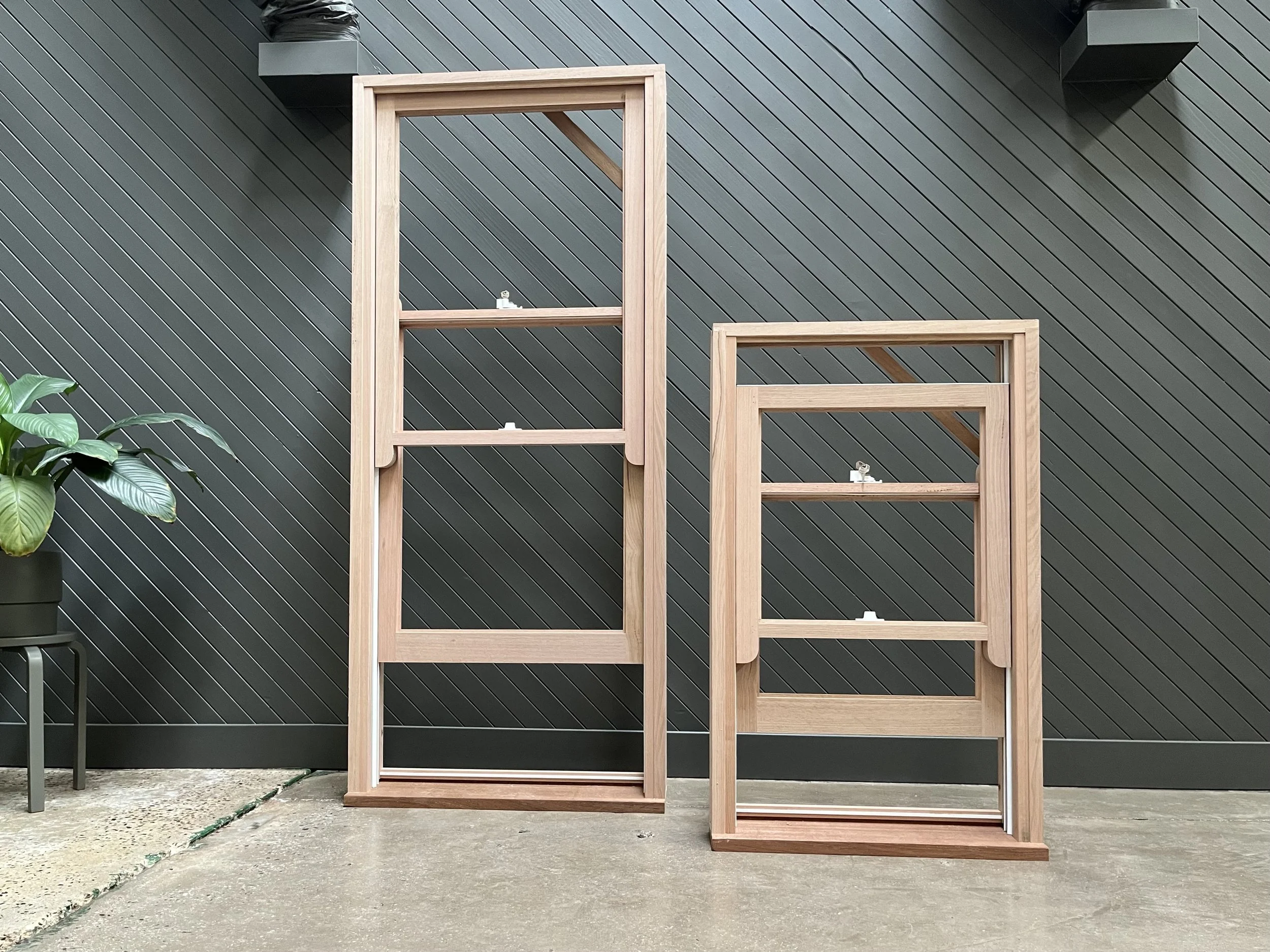 Two wooden plant stands of different heights placed on a concrete floor against a dark gray slatted wall. There is a potted plant with broad green leaves on a small black table to the left, and on the top shelves of the plant stands, there are small 