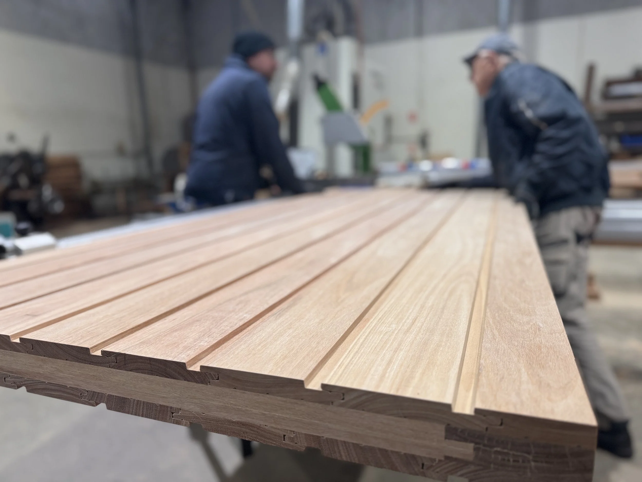 Wooden planks on a workbench in a woodworking shop with two workers in the background.
