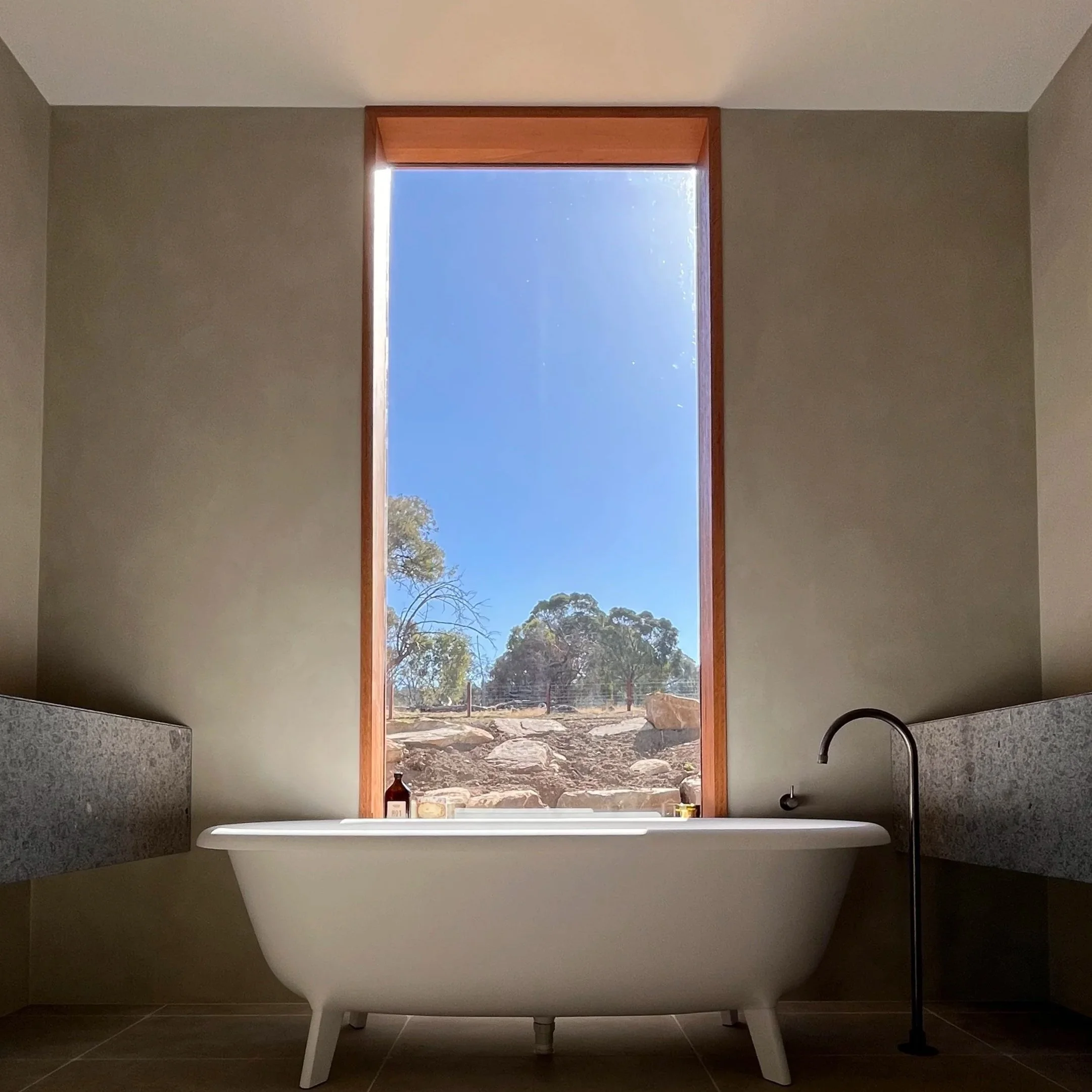 Empty bathtub in a modern bathroom with a large window showing an outdoor landscape with rocks and trees, and a clear blue sky.