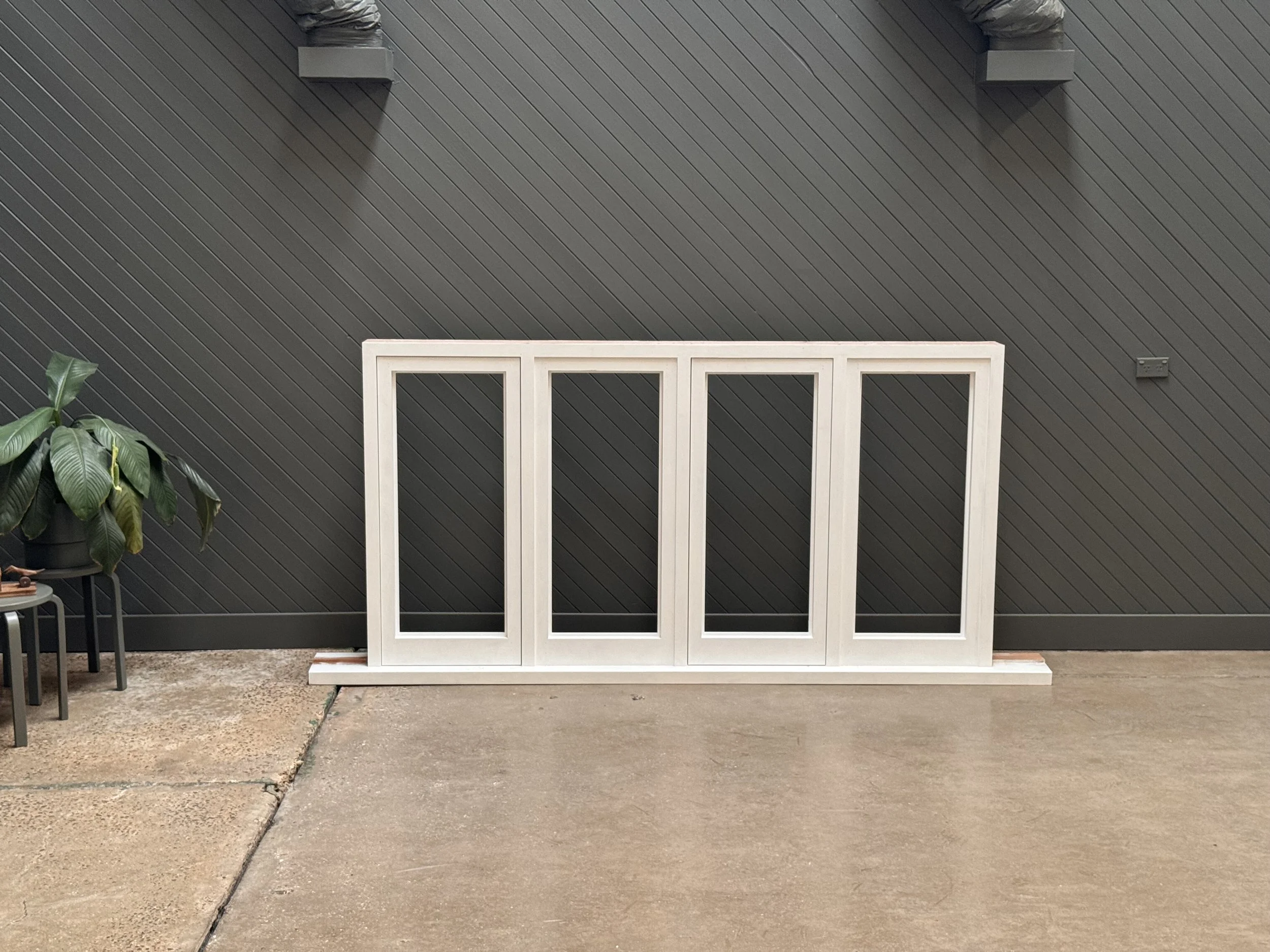 A white wooden frame with four vertical sections leaning against a dark gray wall with diagonal paneling, next to a potted plant on a small black table, on a concrete floor.