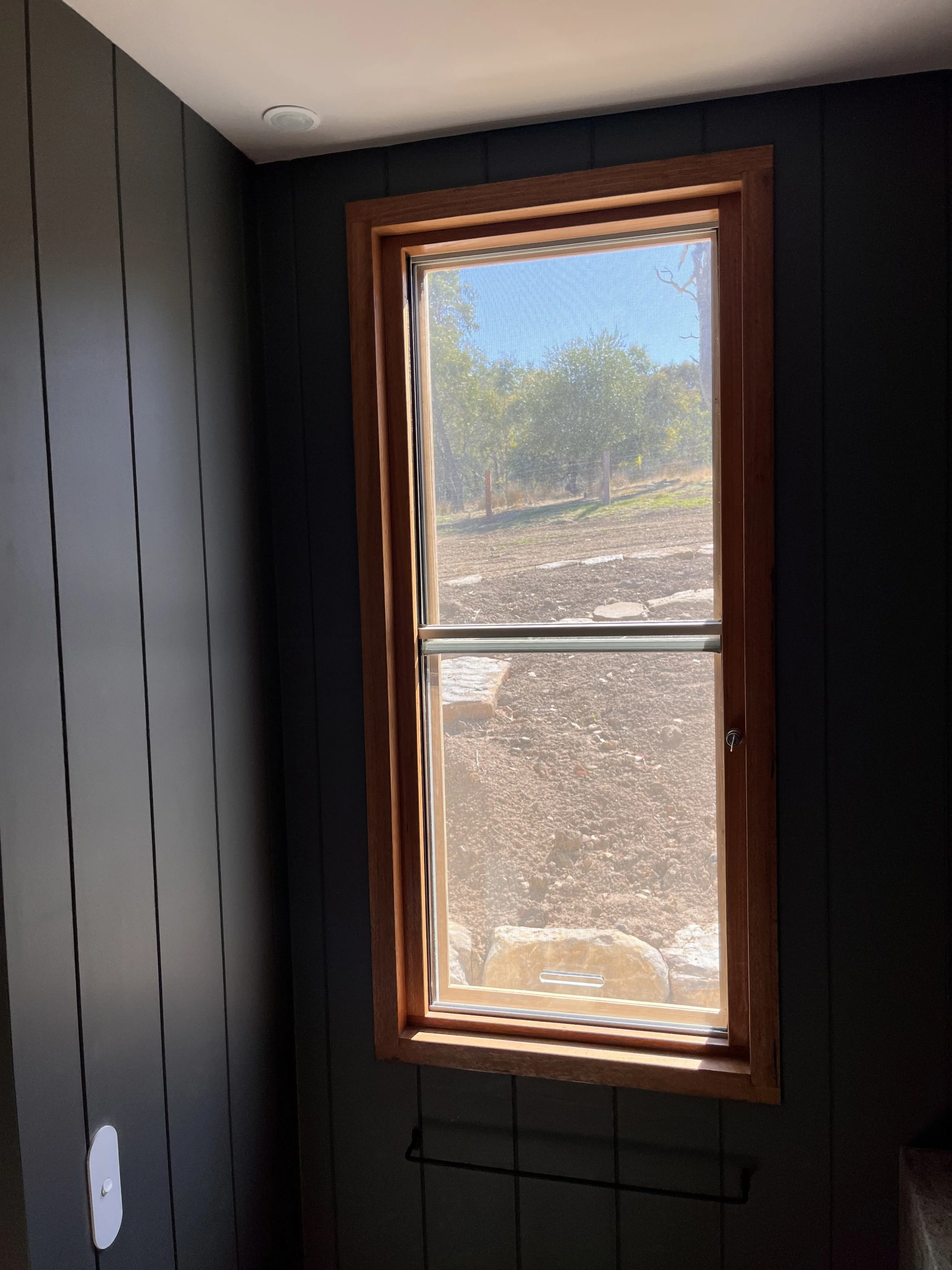 A window with a wooden frame looking out onto a dirt landscape with some trees and a blue sky.