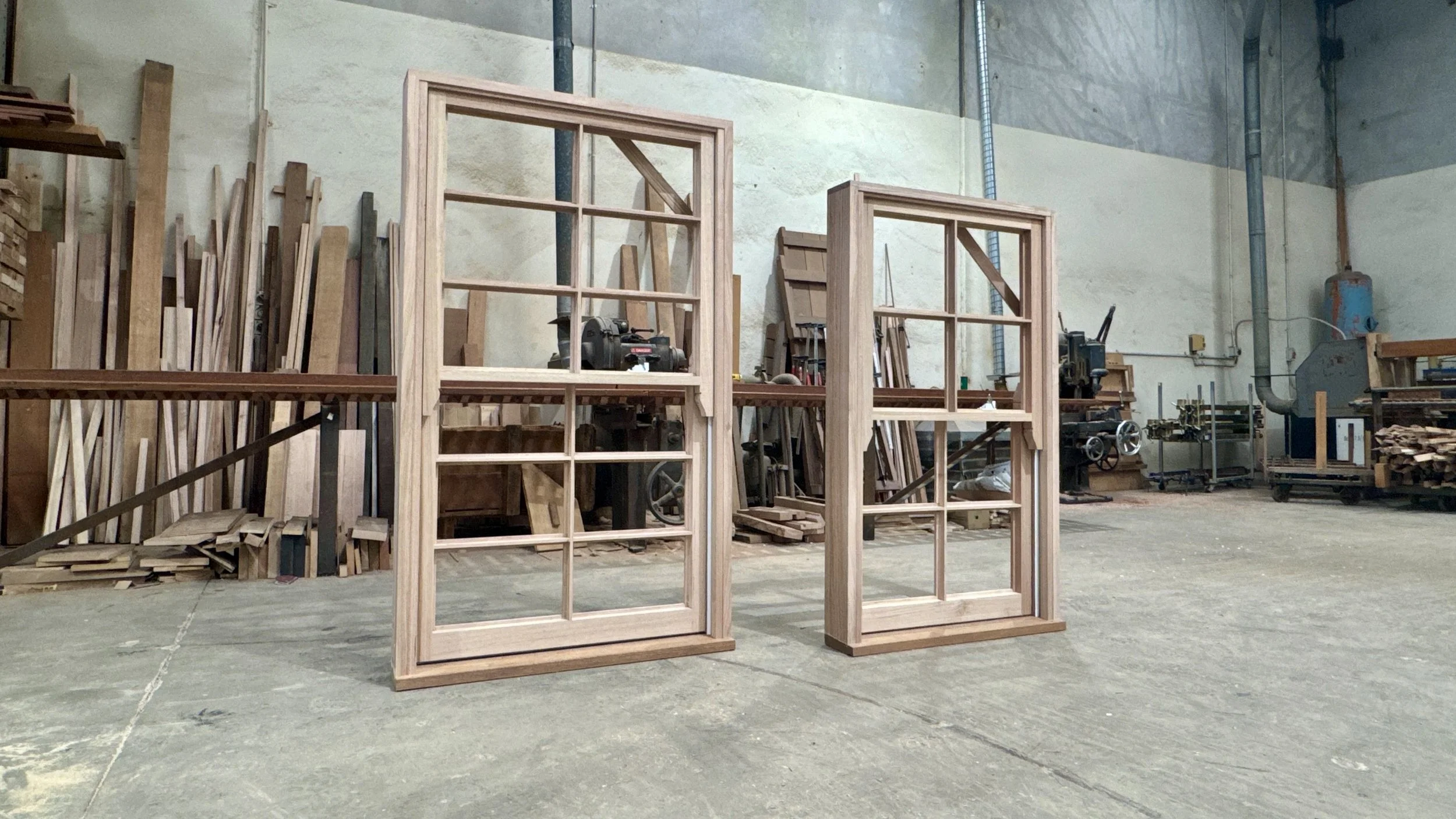 Two unfinished wooden window frames with grid panes are placed on a concrete floor in a woodworking shop. The background contains shelves of wood, tools, and machinery.