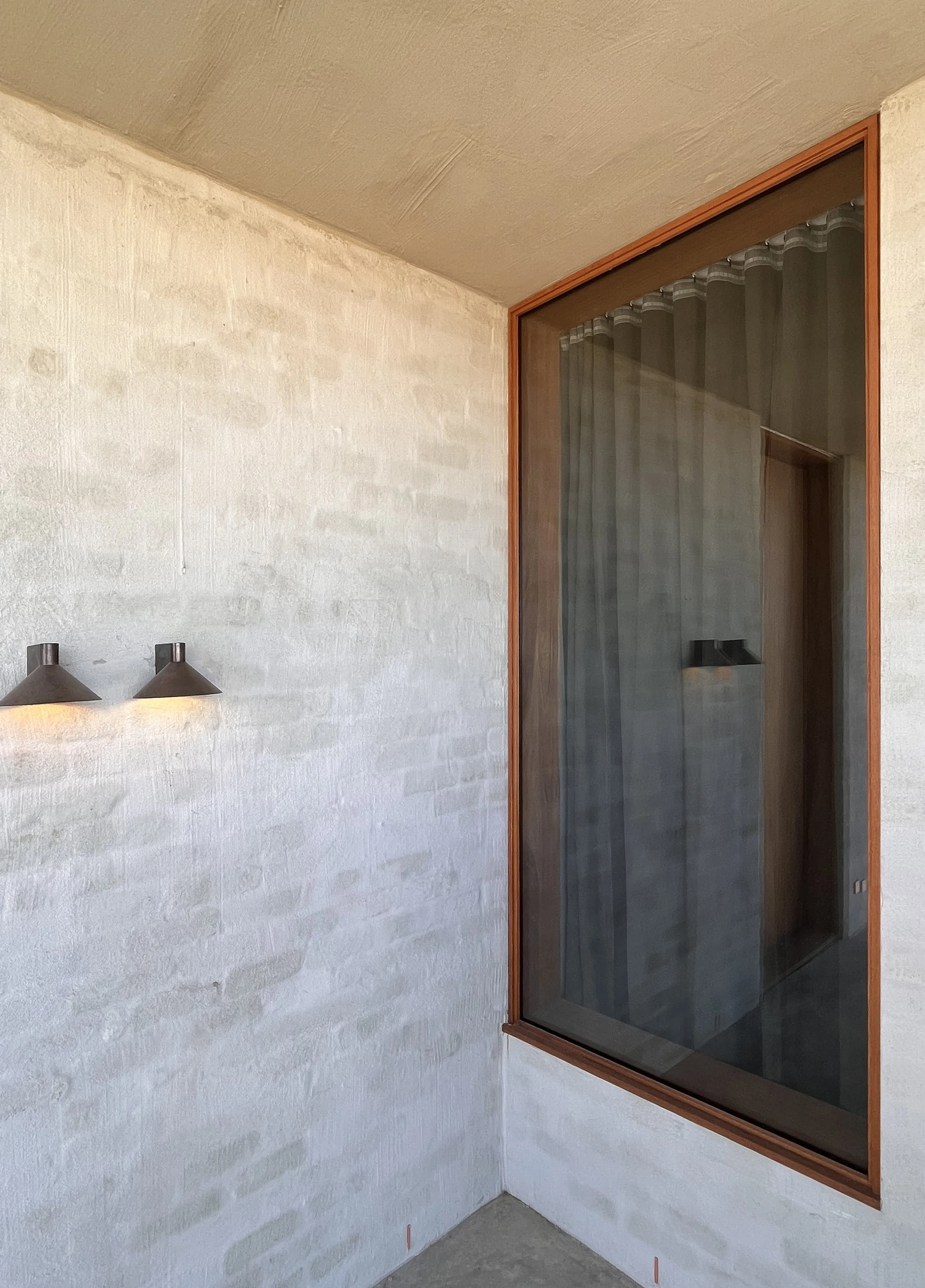 Corner of an outdoor porch with a textured white wall, two modern wall lights, and a large window with wooden frame, revealing a curtain and part of an interior shower space.