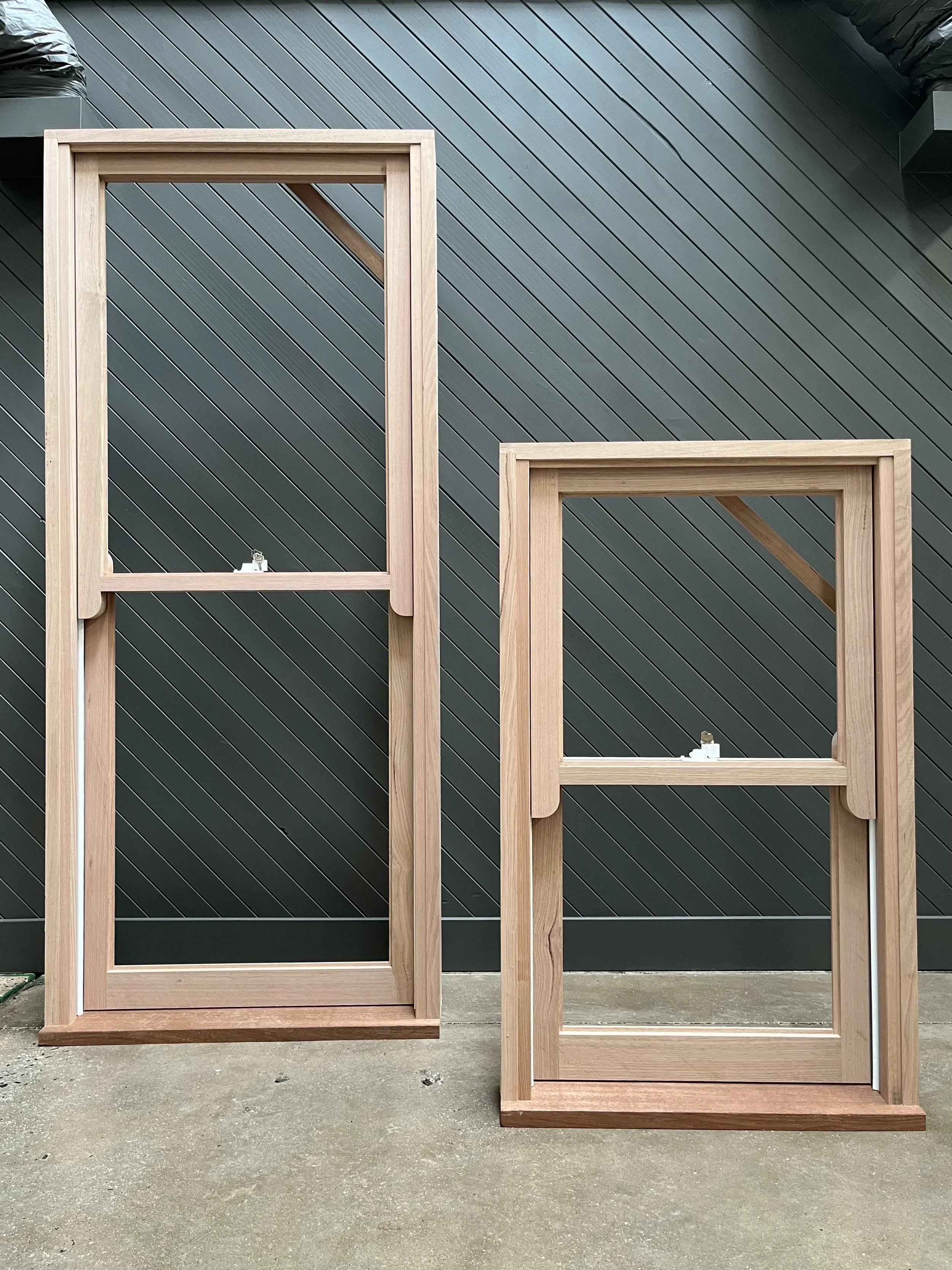 Two wooden screen frames with no screens, placed outdoors against a dark gray diagonal wooden wall, on a concrete ground.