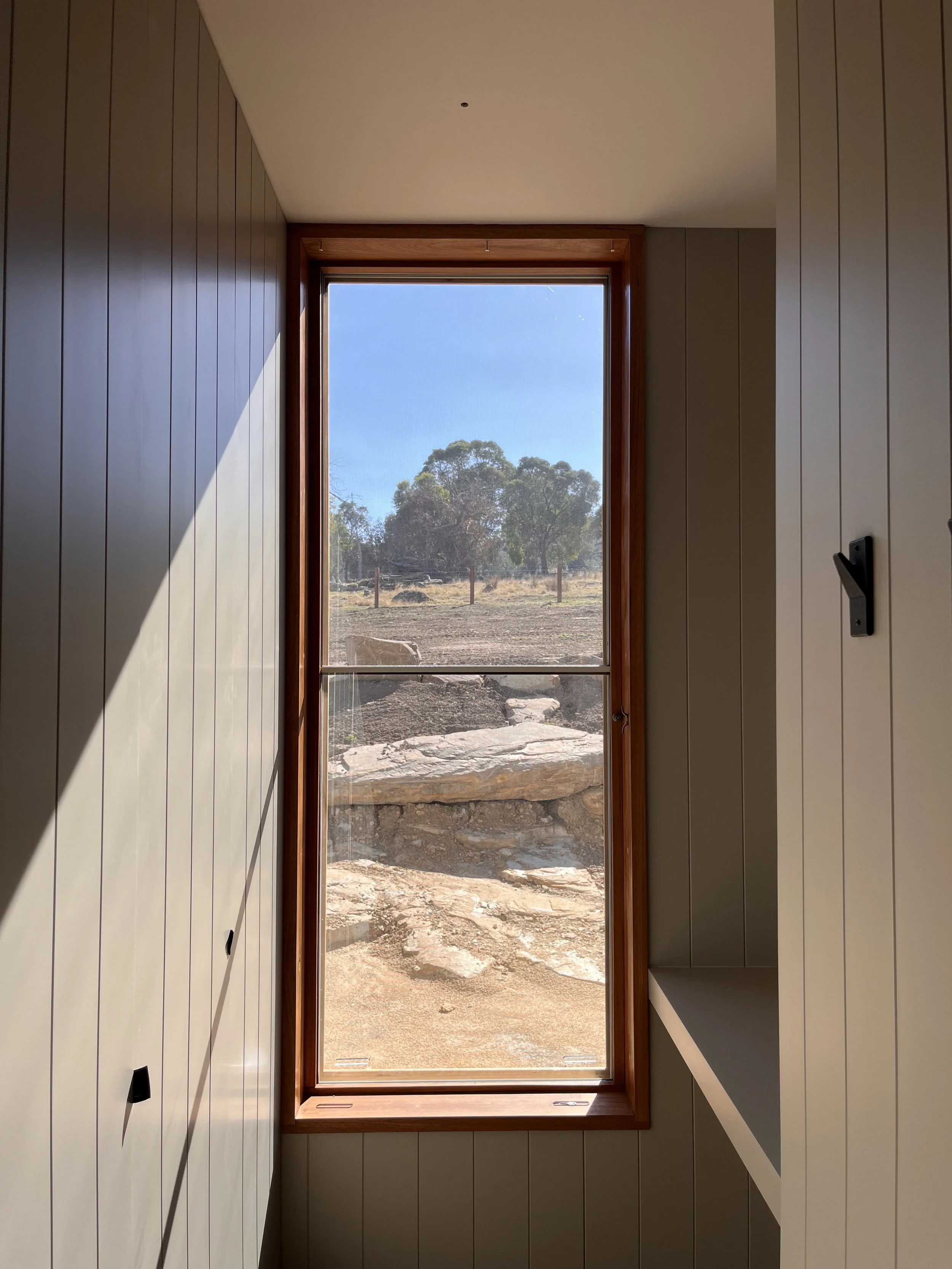 Interior view of a room with vertical wood-paneled walls, a large window with a wooden frame, and a landscape with rocks, trees, and a clear blue sky outside.