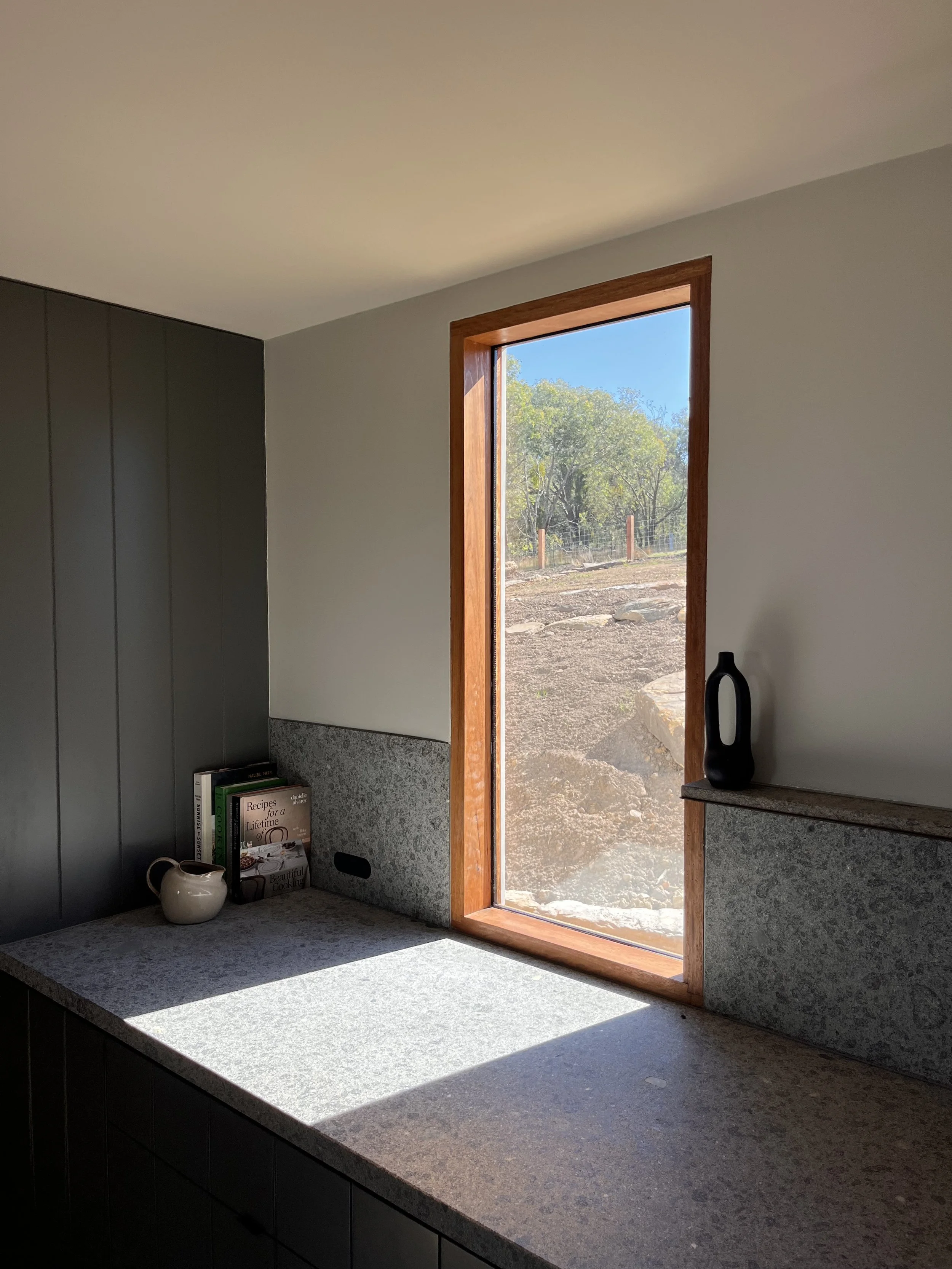 A minimalist interior kitchen scene with a granite countertop, a small white jug, a few books, and a black vase. A large wooden-framed window shows an outdoors view of trees and dirt ground, letting natural light into the room.