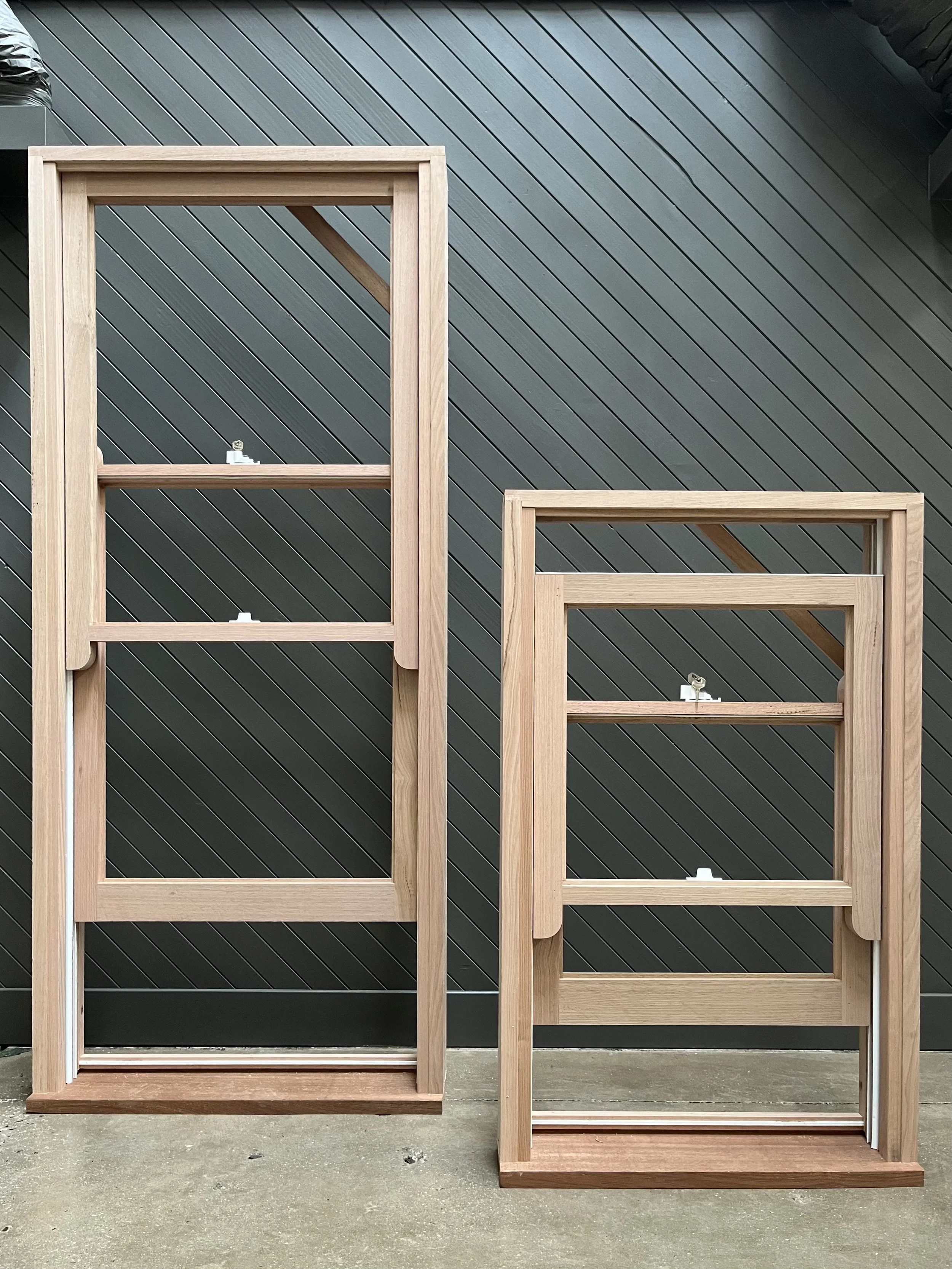 Two unfinished wooden framed beds or shelves with a light wood color, placed on a concrete floor against a dark gray, diagonally panelled wall.