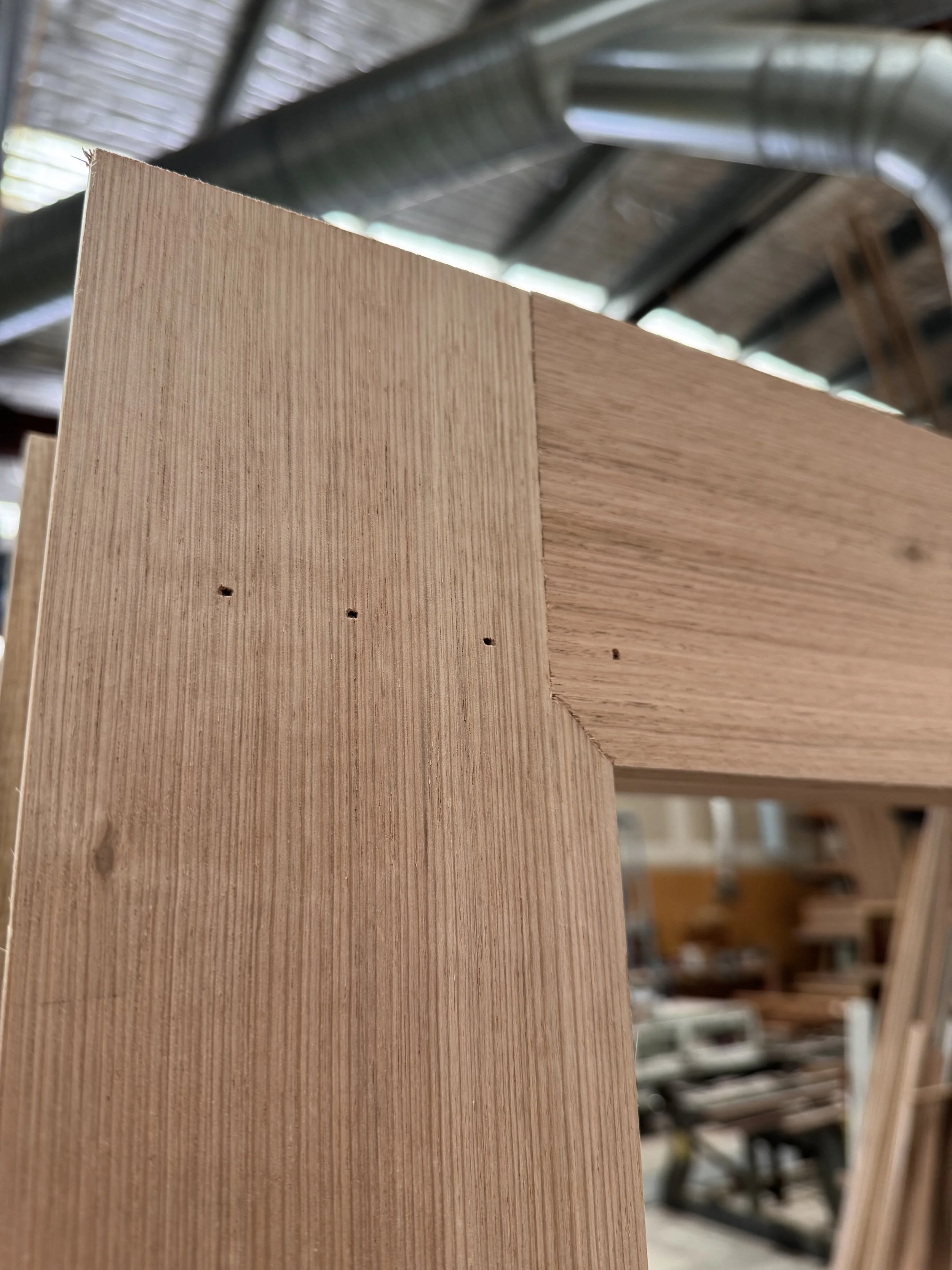 Close-up of a wooden corner joint with visible nail holes, in a woodworking shop with tools and wood in the background.