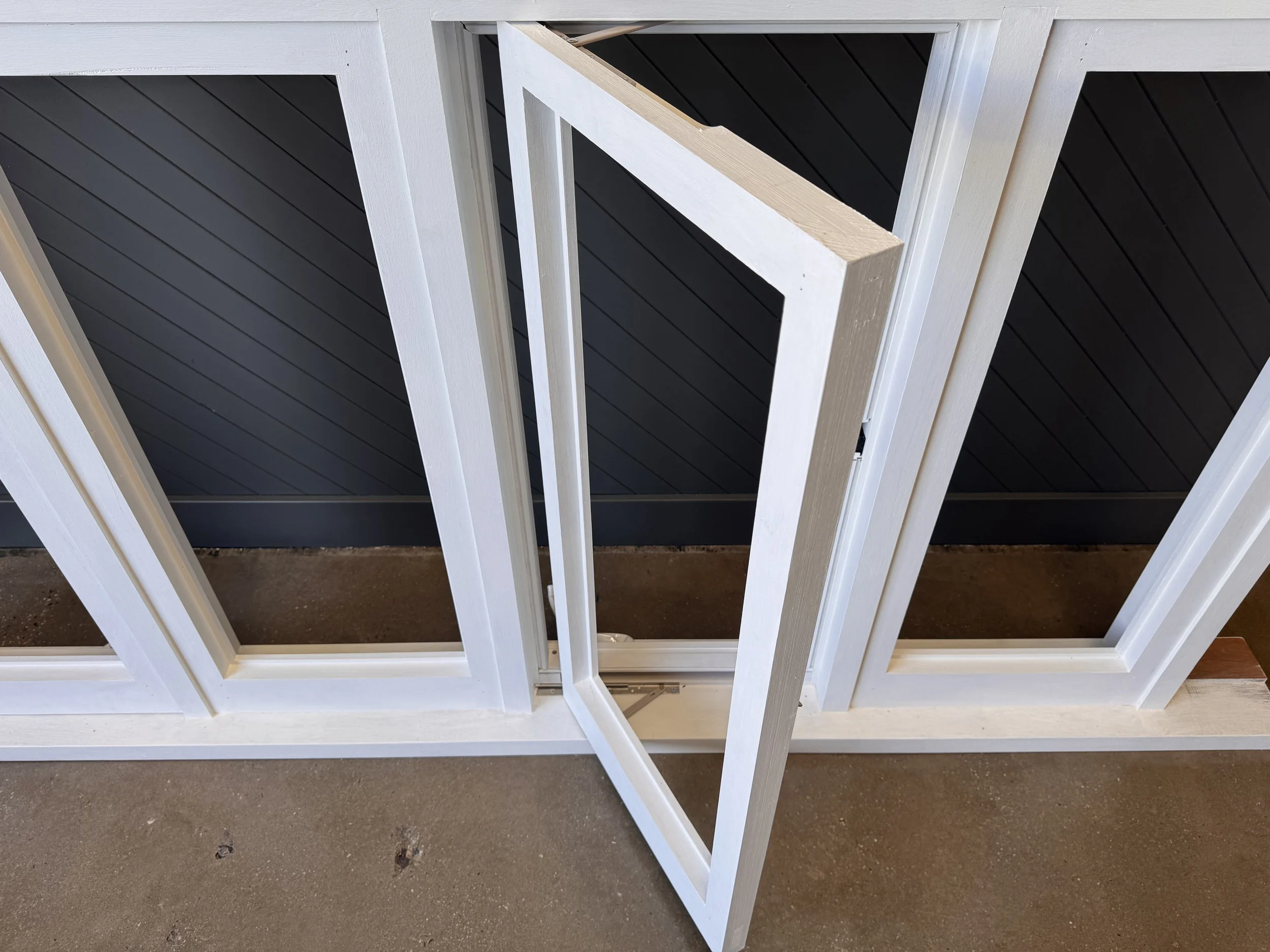 Close-up of a newly installed window with white framing, partially open, overlooking a black exterior wall and a concrete floor.