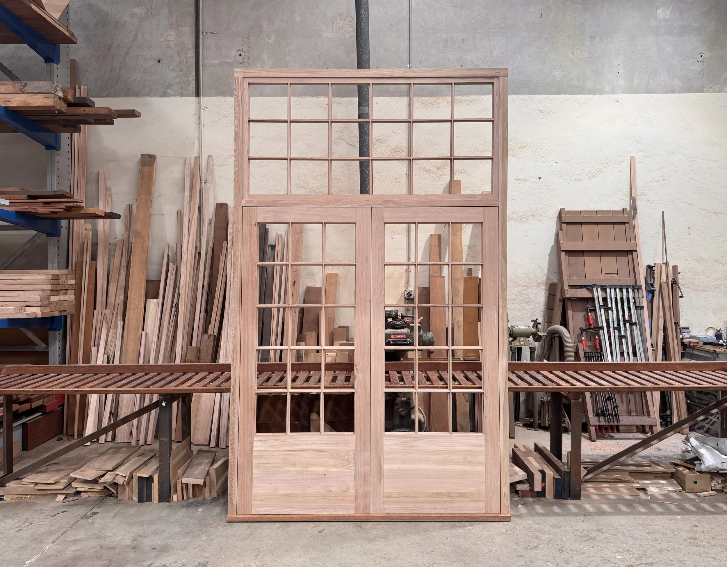 unfinished wooden doors with glass panes leaning against a work table in a woodworking shop with various wood pieces and tools in the background.