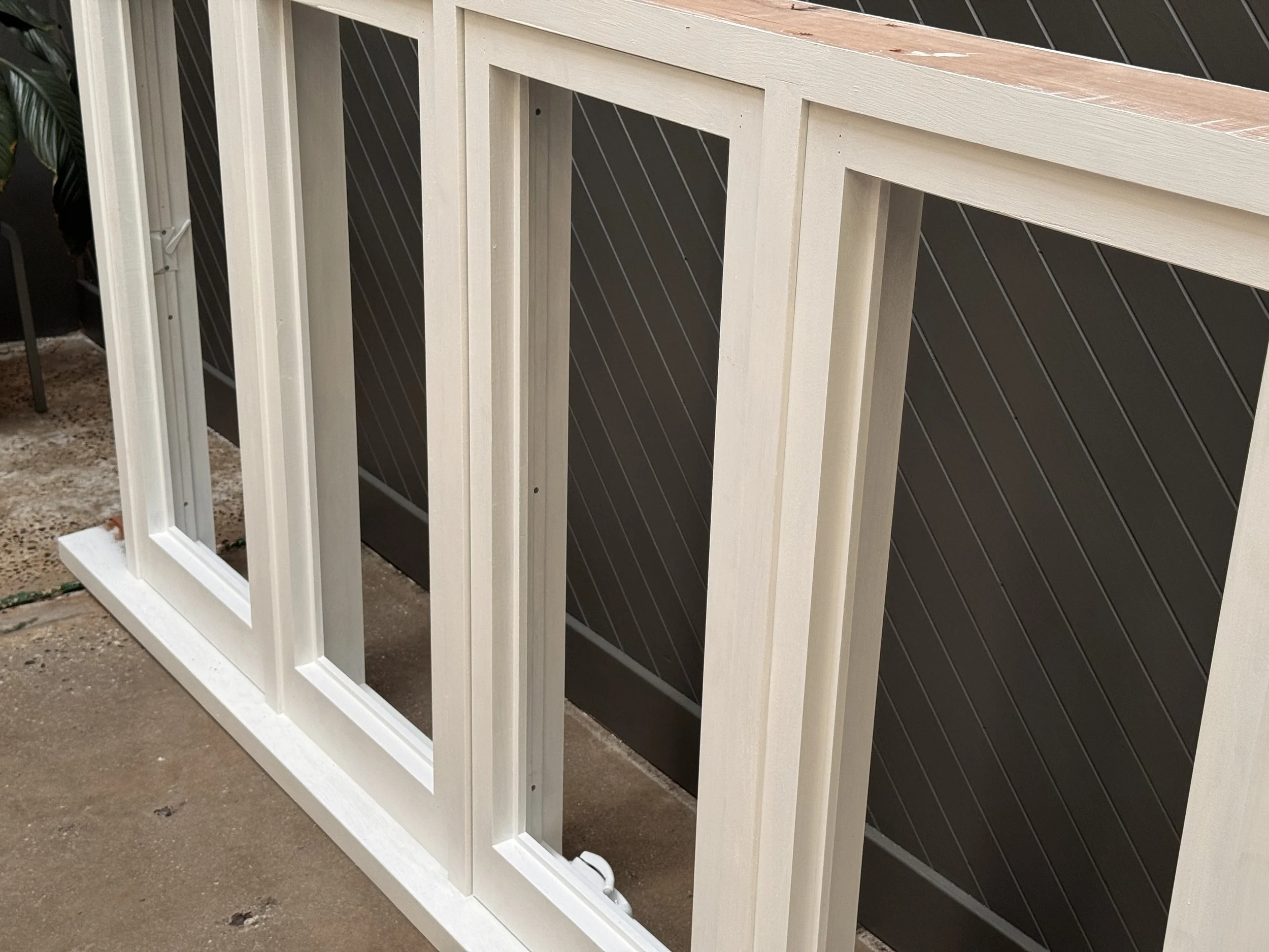 unfinished wooden window frame leaning against a dark brown slatted wall with a plant and chair to the side, on a concrete floor