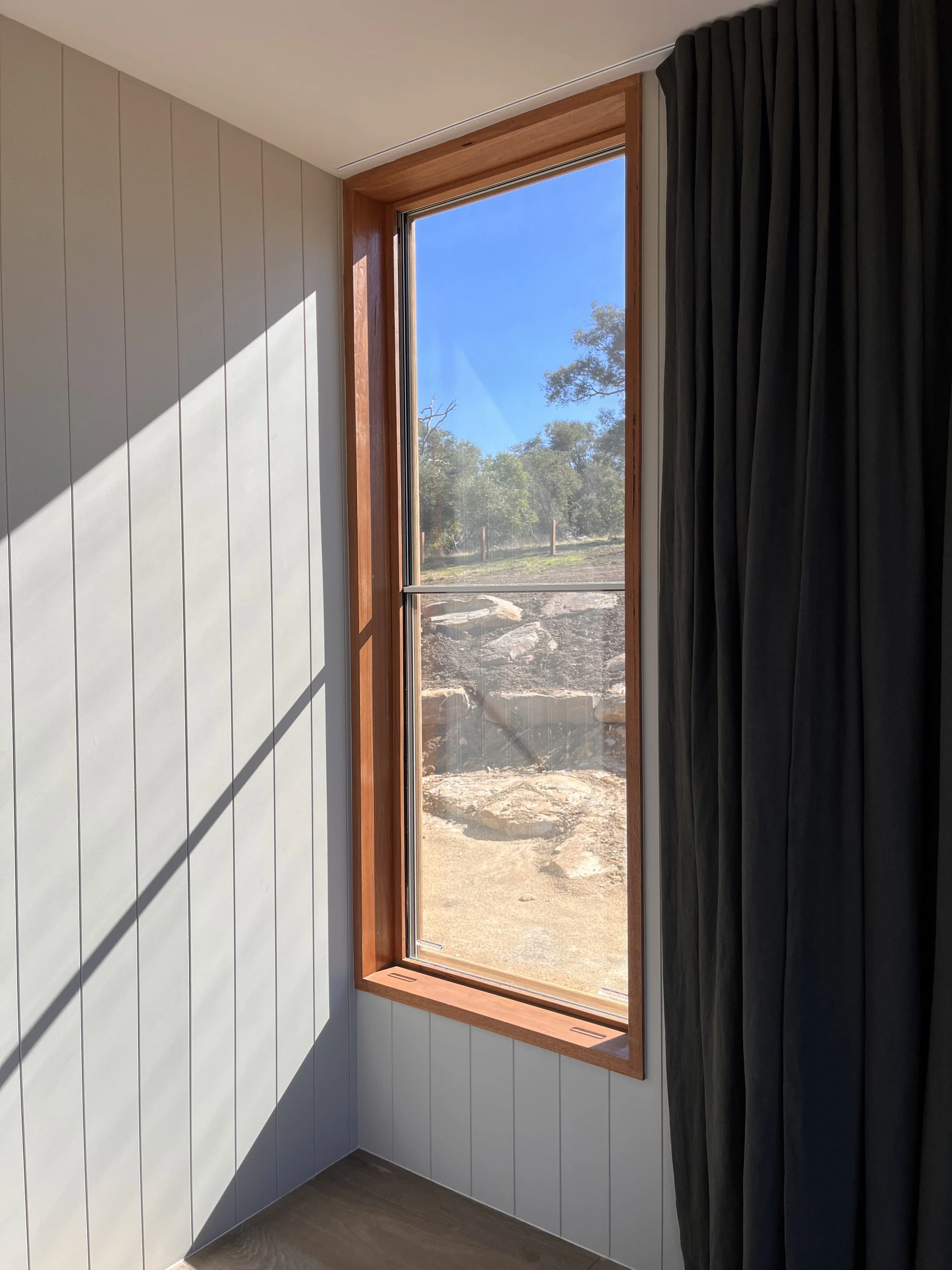 Interior of a room with a large window framed in wood, showing a view of trees and rocks outside. The room has light-colored paneling on the walls and a dark curtain partially covering the window.