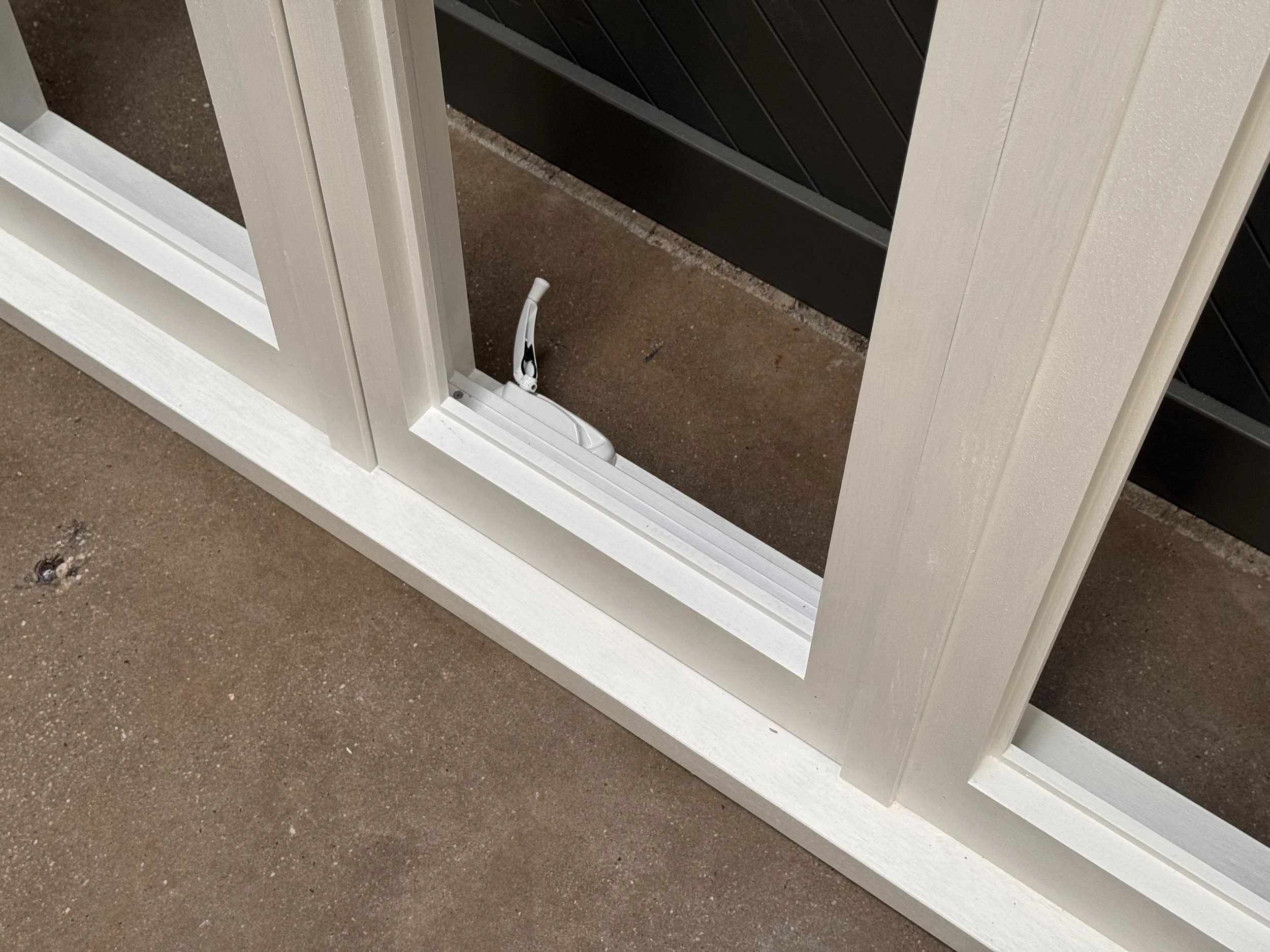 Close-up of a white screen door frame with a broken latch at the bottom, on a concrete surface.