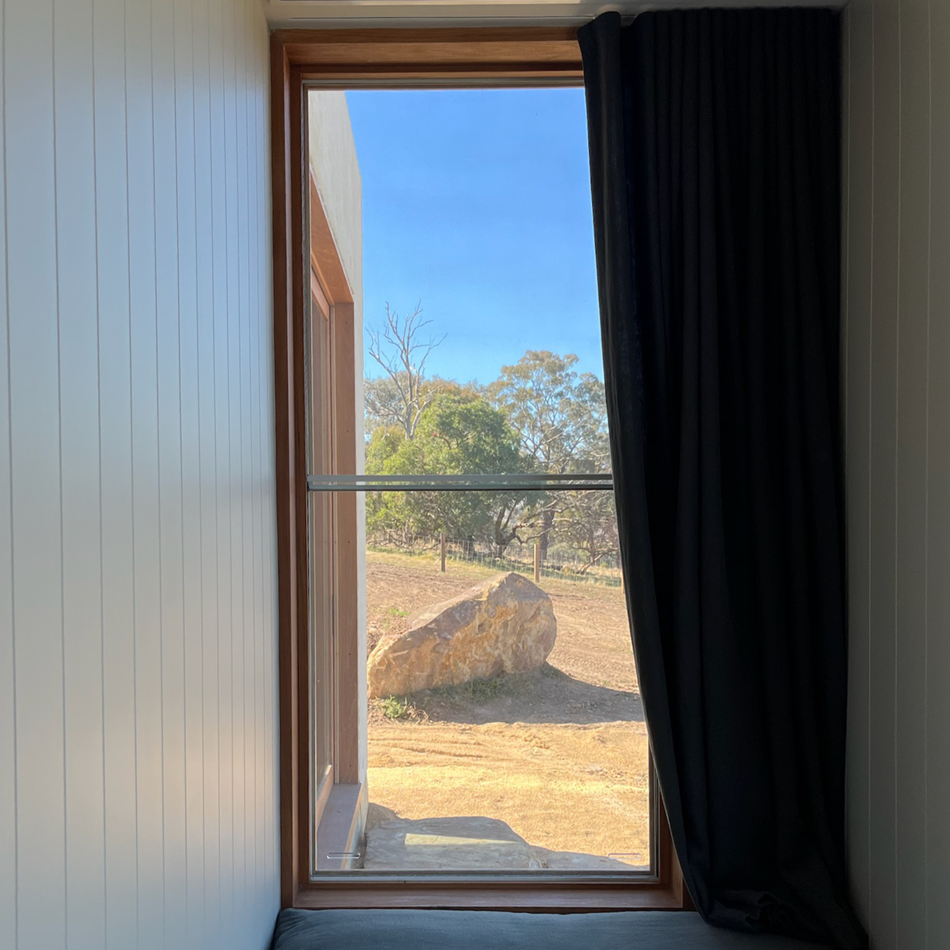 Interior view of a window with a black curtain pulled to the side, revealing a landscape with a large rock, trees, and a clear blue sky.