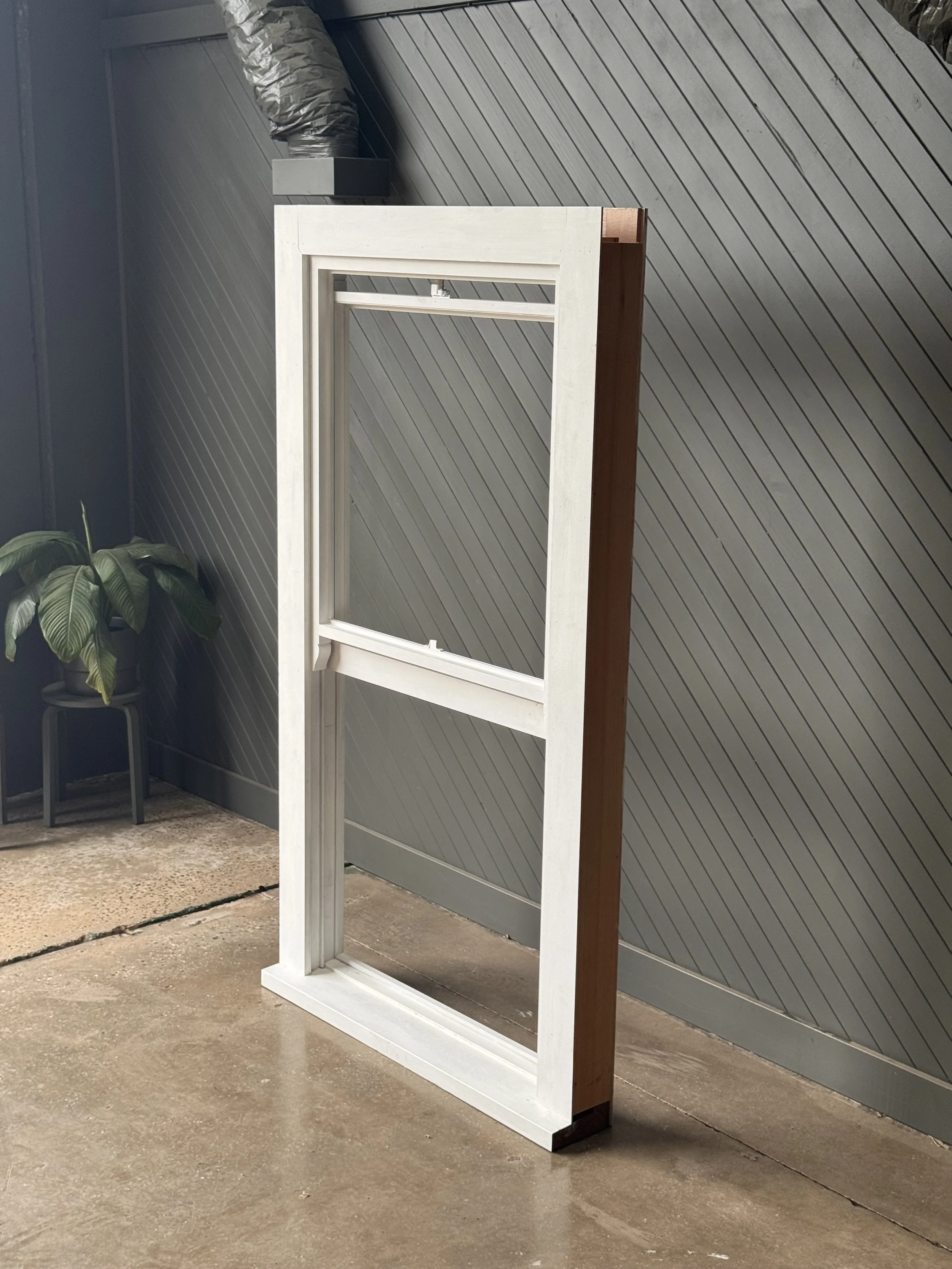 Unassembled white window frame leaning against a beige wall with a small potted plant nearby and an industrial vent pipe on the ceiling.