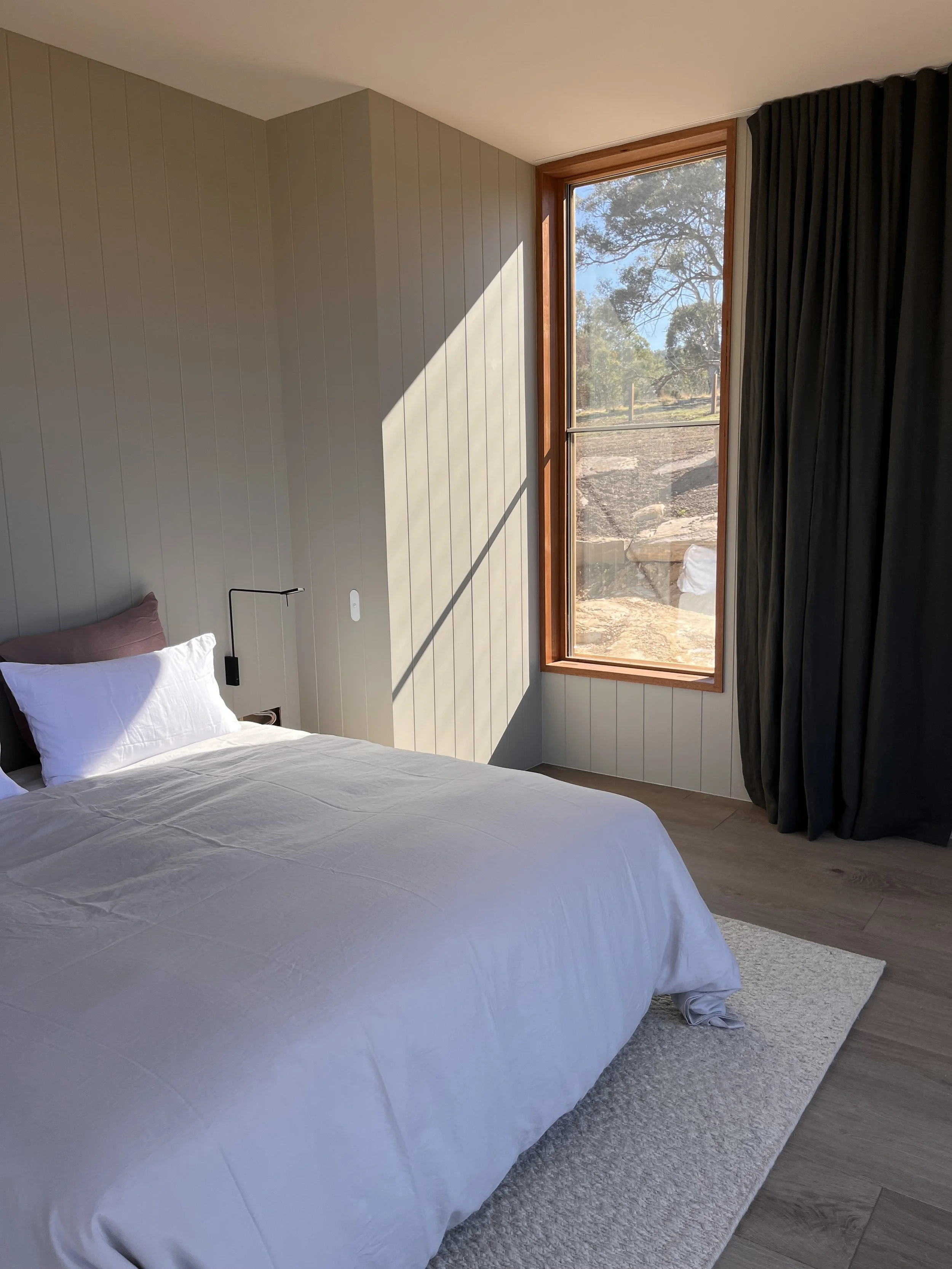 A neatly made bed with white linens and pillows in a sunlit bedroom with wood-paneled walls, a large window, and a view of trees outside.