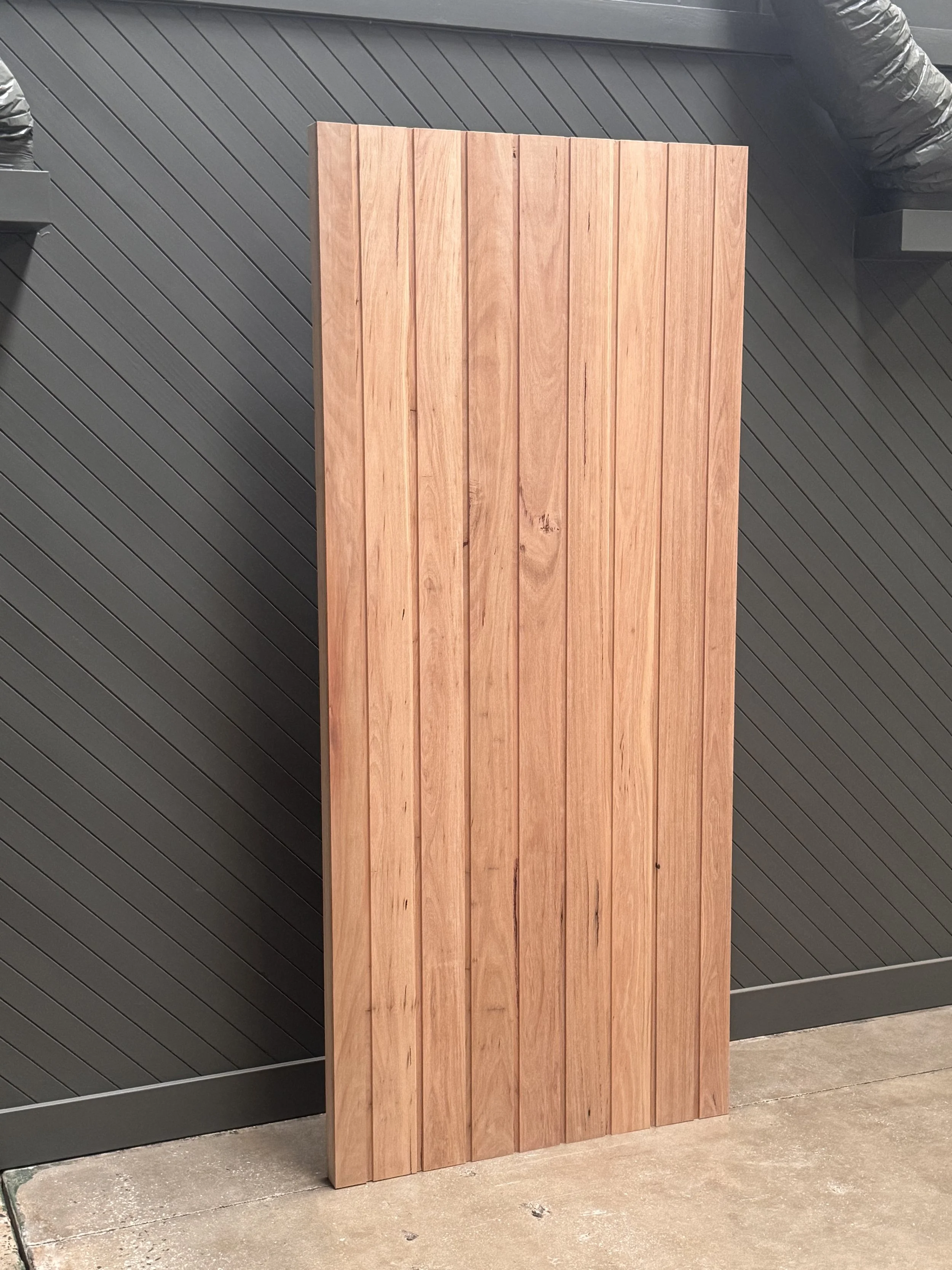 Vertical wooden panel leaning against a dark gray wall on a concrete ground.