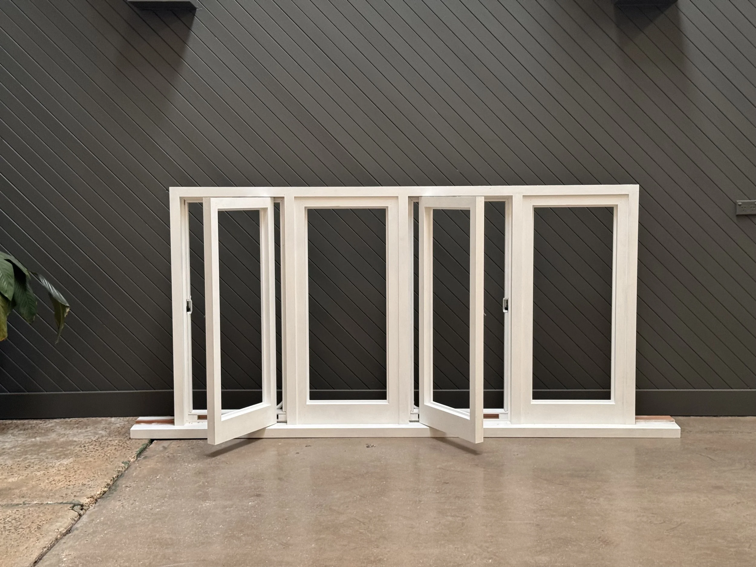 Unassembled white window frame with three sections leaning against a dark wall on a concrete floor.