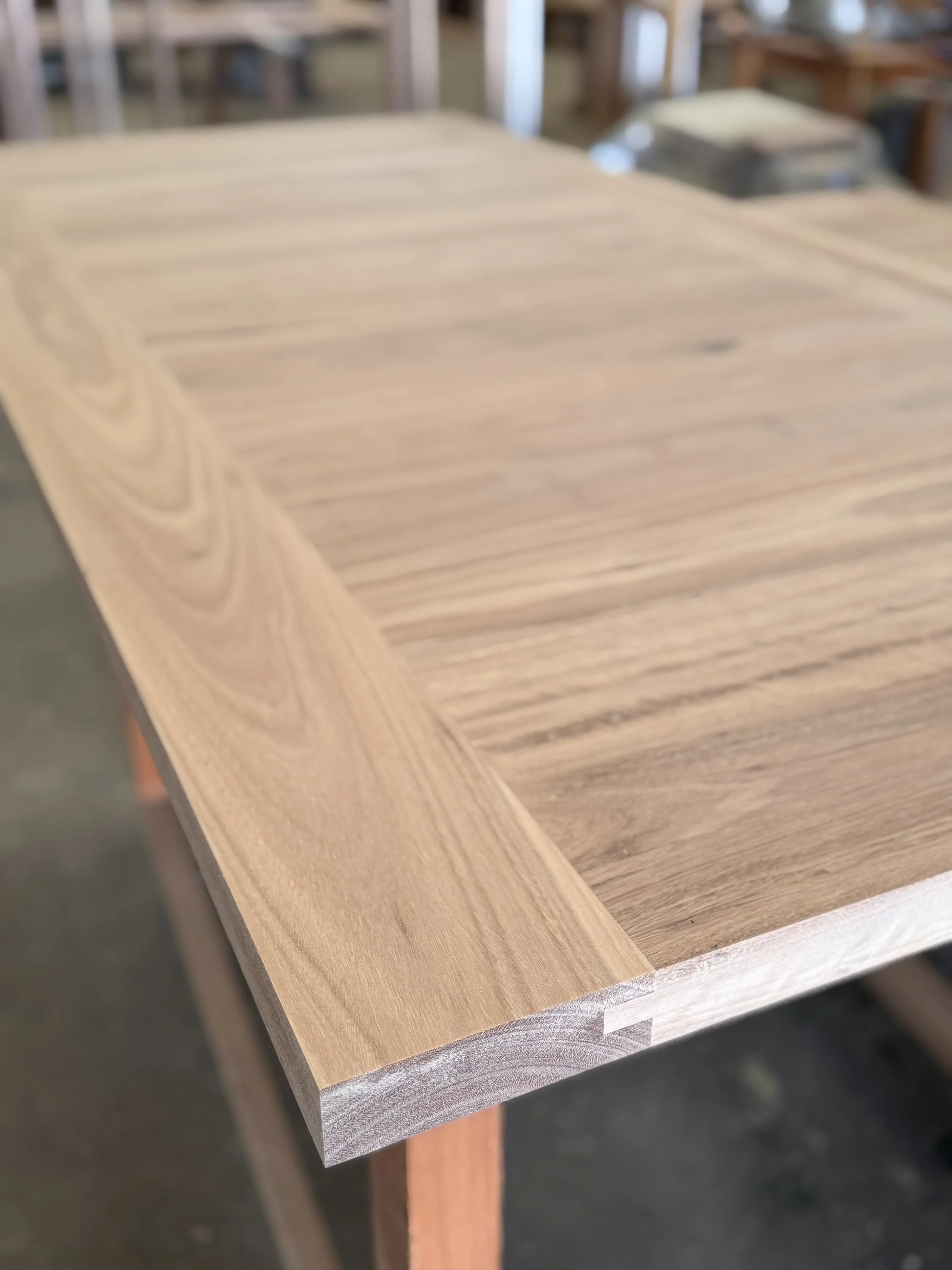 Close-up of a wooden table with a smooth, finished surface and visible wood grain, with additional furniture in the background.