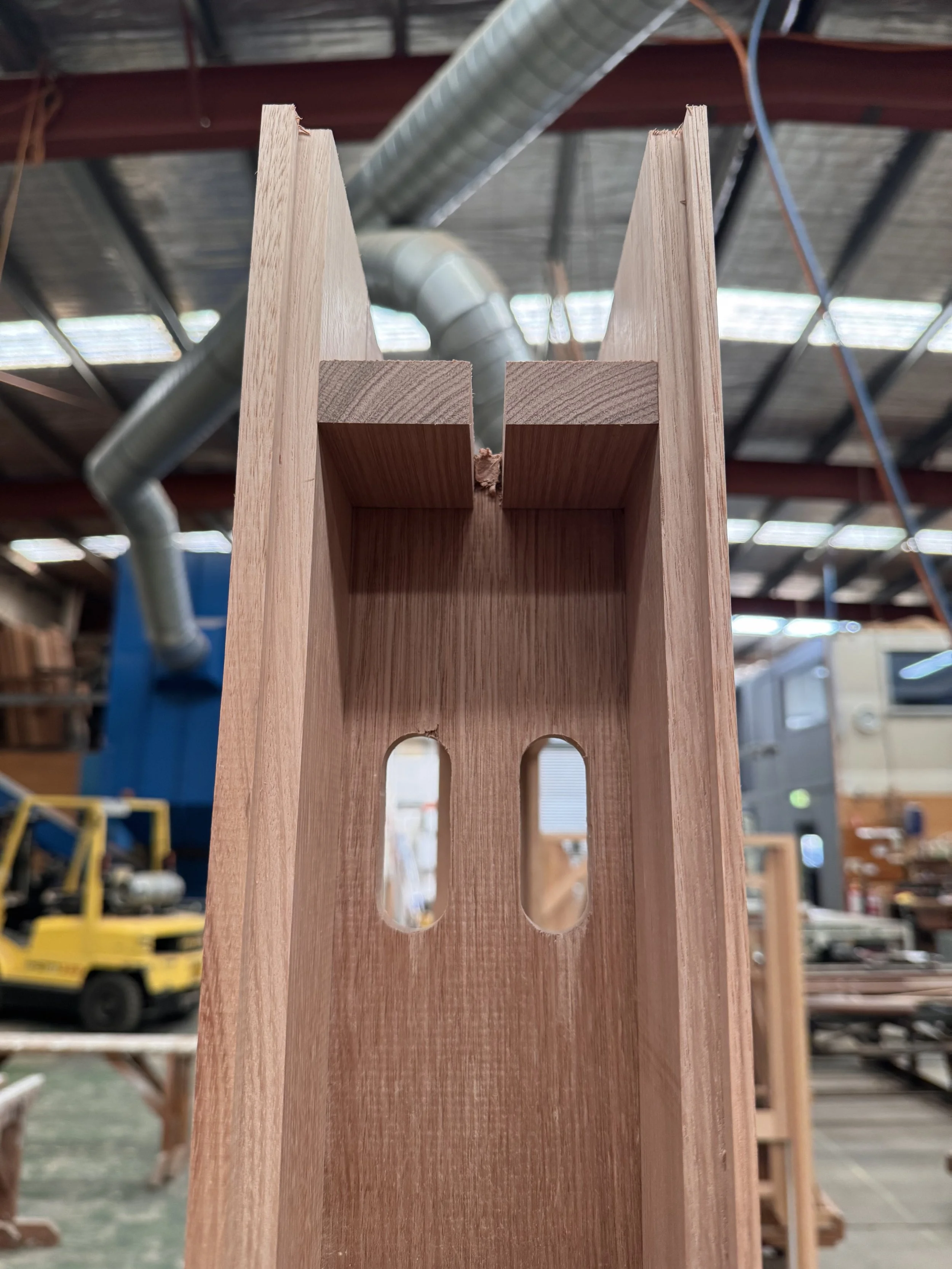 Close-up view of a wooden piece with precise cut-outs, likely part of furniture construction, inside a workshop with industrial elements in the background.