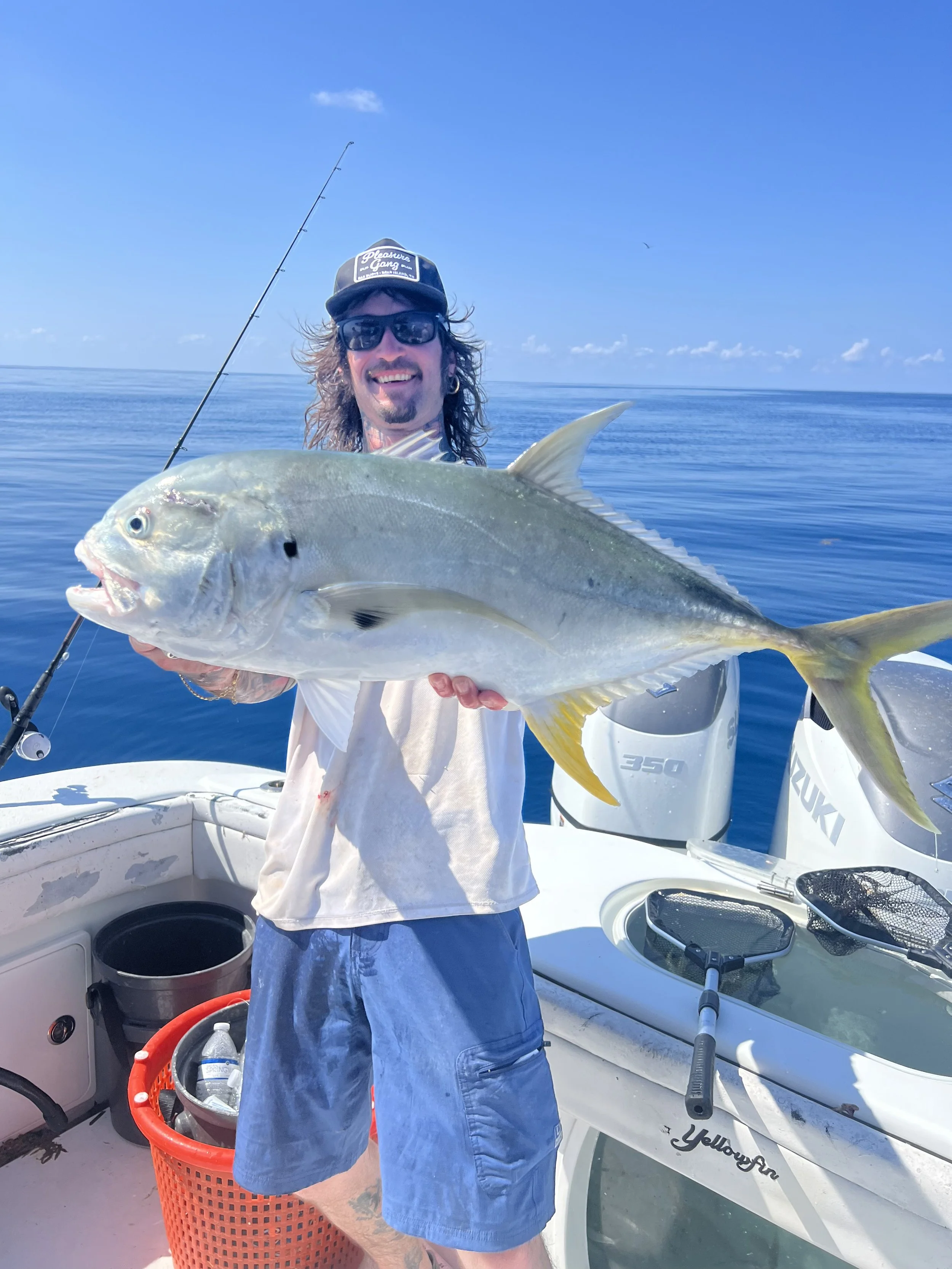 A man with long hair, wearing sunglasses, a black cap, a white shirt, and blue shorts, holding a large fish on a boat in the ocean under a clear blue sky.