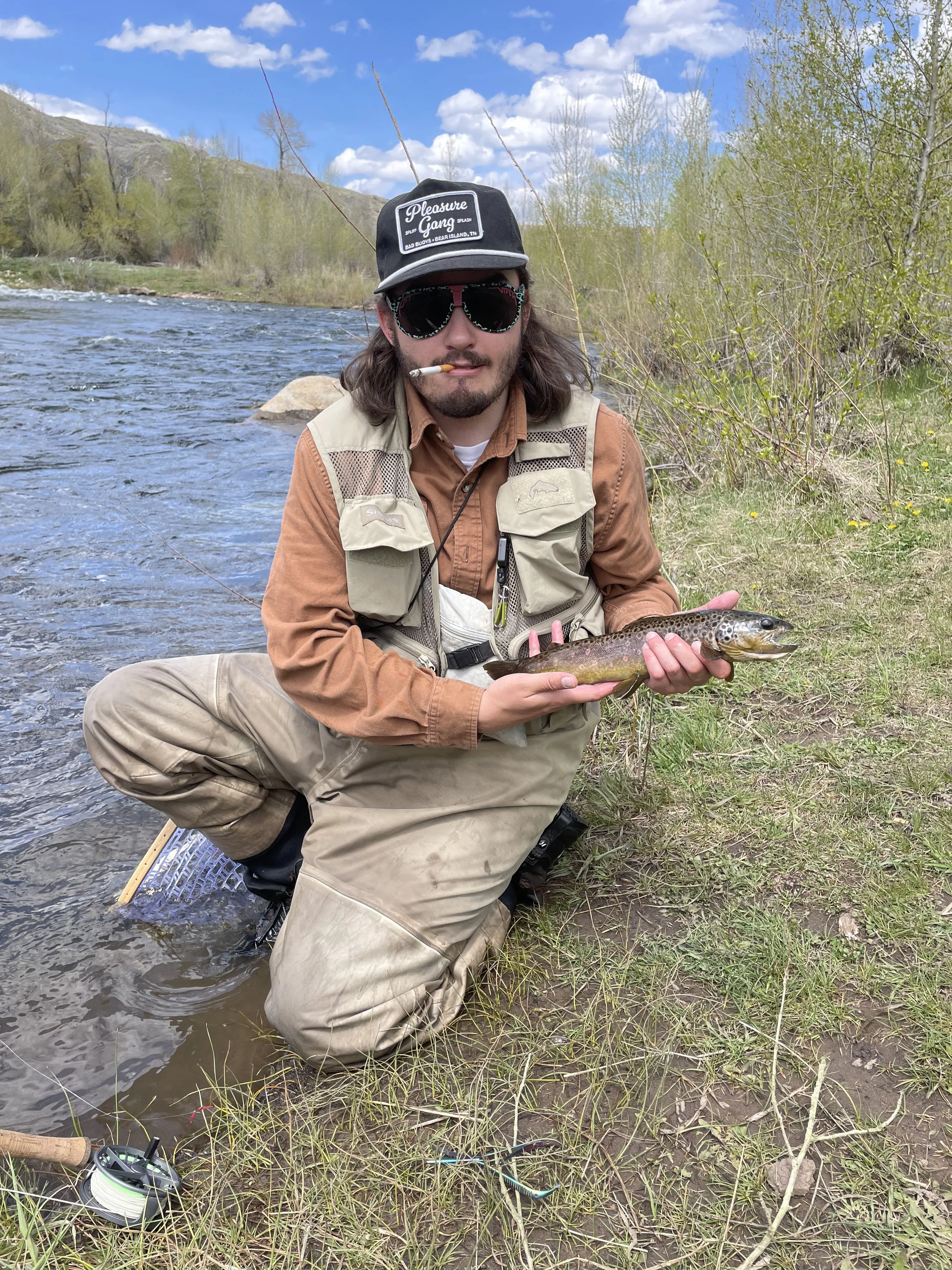 A man kneeling near a river holding a freshly caught fish, wearing sunglasses, a cap, and outdoor fishing gear with the river and trees in the background.